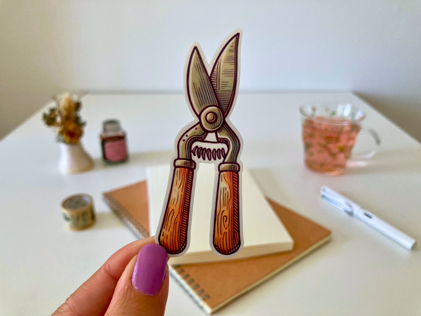 A hand with purple nail polish holds a cartoon sticker of vintage-style garden secateurs with wooden handles. The background features a desk with a notebook, pen, dried flowers, washi tape, a jar, and a glass cup of tea.