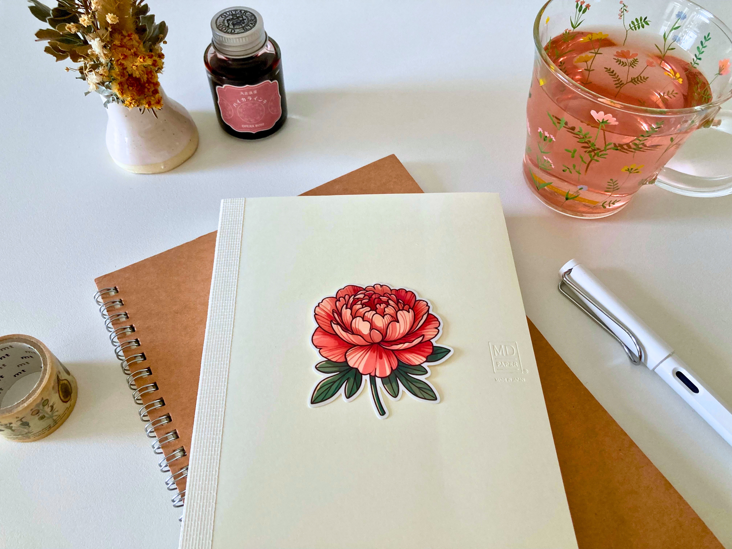 A cartoon peony sticker with layered red and pink petals and green leaves is placed on a cream-colored Midori journal. Surrounding items include a floral glass mug with pink tea, a white pen, a brown notebook, washi tape, dried flowers, and a bottle of ink.