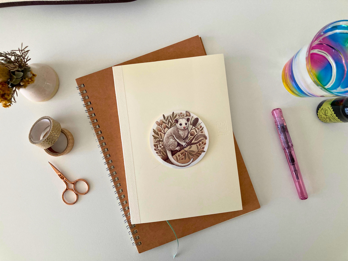 A round Australian possum sticker sits on a cream-colored notebook, which rests on a brown spiral-bound journal. The sticker features a fluffy possum perched on a branch surrounded by native botanicals in earthy tones. Around the notebooks are a pink fountain pen, gold scissors, washi tape, a glass with rainbow swirls, dried flowers in a vase, and an ink bottle. The cozy desk setup highlights the waterproof, scratch-resistant sticker, perfect for decorating journals, laptops, and more.