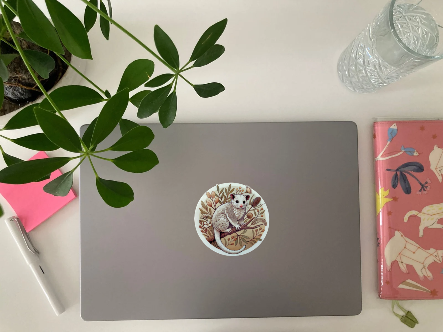 A silver laptop with a round Australian possum sticker sits on a white desk. The sticker features a fluffy possum perched on a branch, surrounded by native botanicals in warm earthy tones. Green plant leaves partially frame the top of the image. Nearby are a pink notebook with animal illustrations, a white fountain pen, pink sticky notes, and a glass of water. The waterproof, scratch-resistant sticker adds a cozy, nature-inspired touch to the workspace.