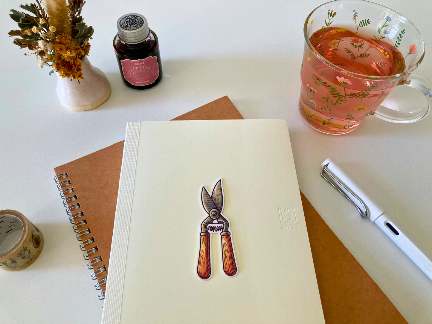 A cartoon sticker of vintage-style pruning shears with wooden handles is placed on a cream-colored Midori notebook, set atop a brown spiral notebook. Nearby are a floral glass mug with pink tea, a white pen, dried flowers in a vase, washi tape, and a small jar of ink.