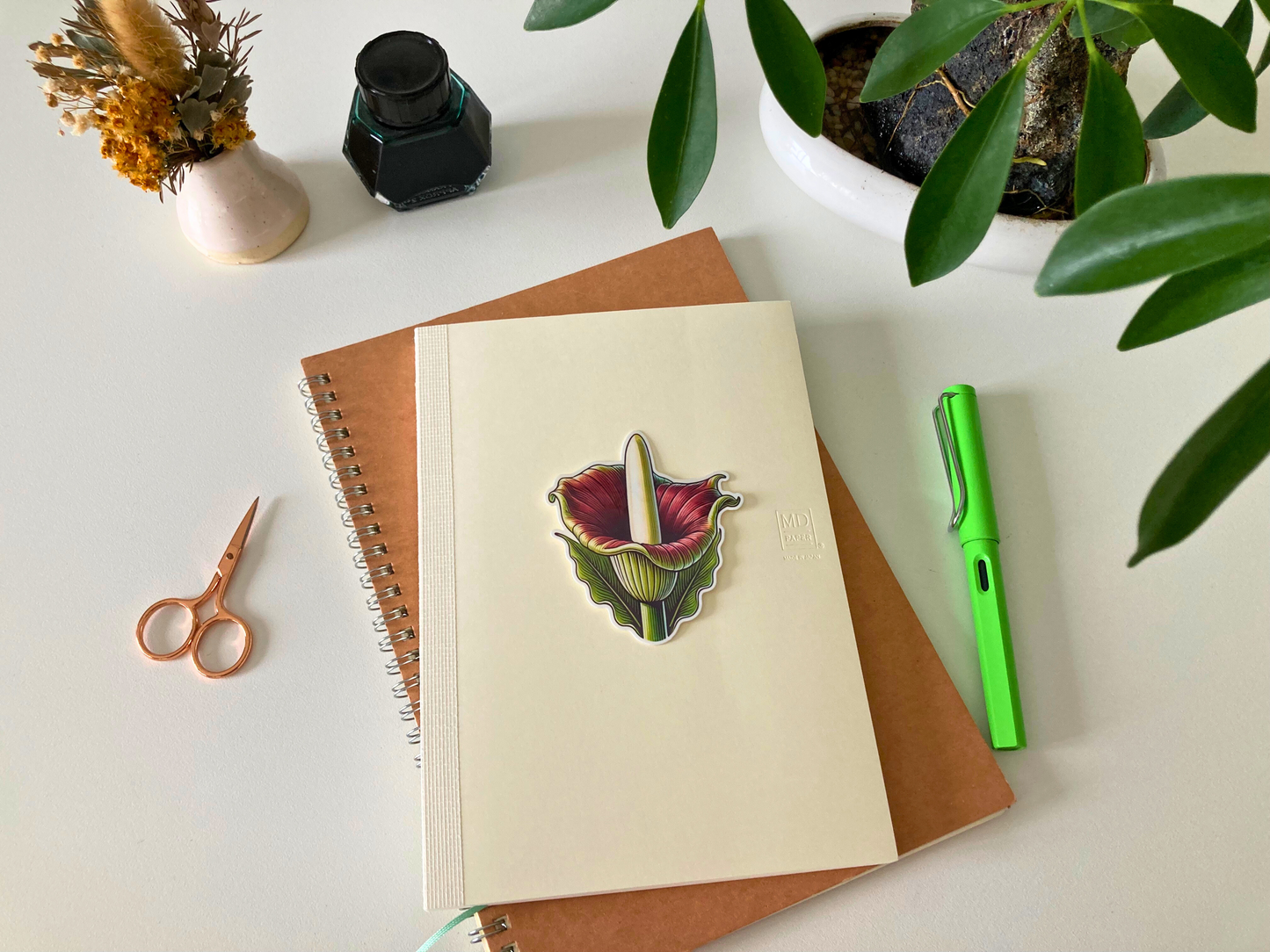 A colorful corpse flower sticker placed on the cover of a Midori journal, surrounded by a green pen, copper scissors, a small vase with dried flowers, and a leafy potted plant.