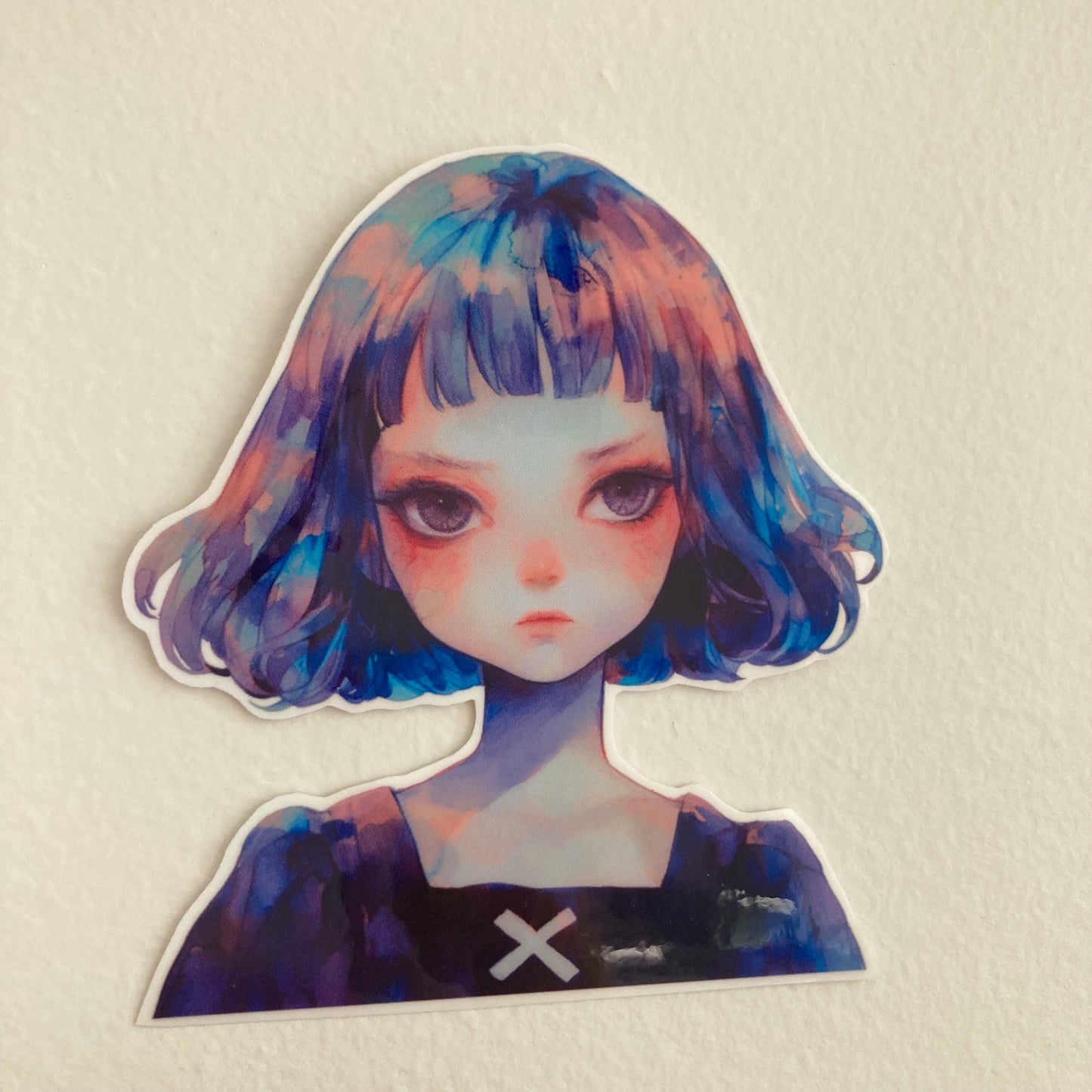 A sticker of a girl with short blue and purple hair, wearing a black top with a white "X," placed against a plain light-colored surface.