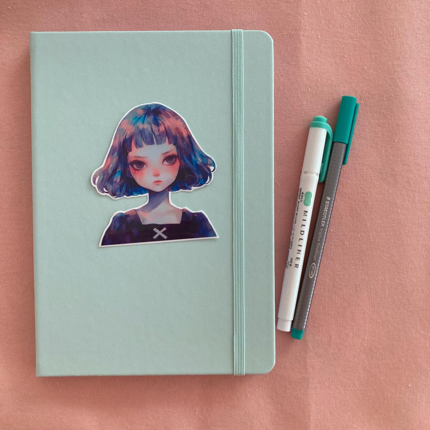 A sticker of a girl with blue and purple hair is placed on a light teal notebook with an elastic band. Next to the notebook are a green marker and a gray pen, all on a soft pink fabric background.