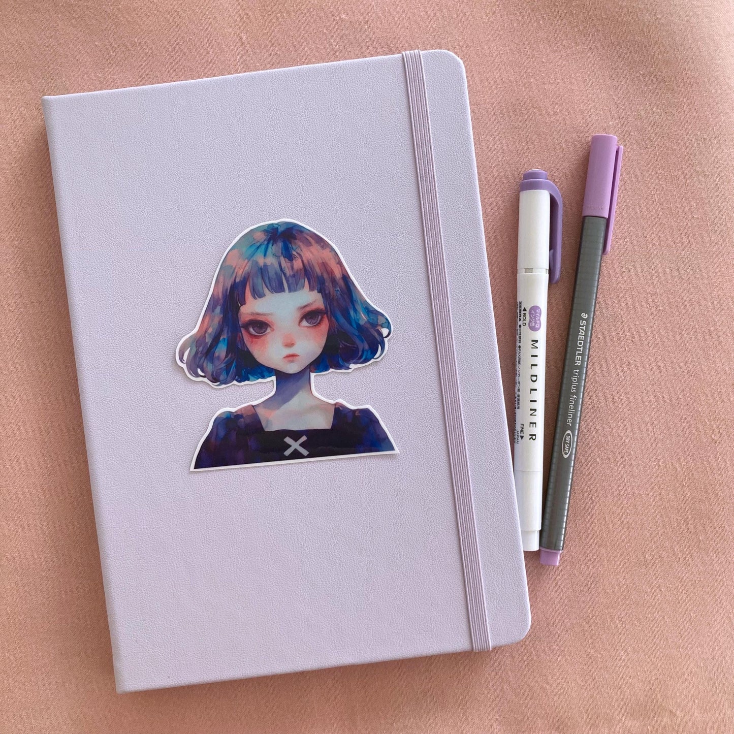 A sticker of a girl with blue and purple hair is on a light lavender notebook with an elastic band. Beside the notebook are a purple marker and a gray pen, all set on a soft pink fabric background.
