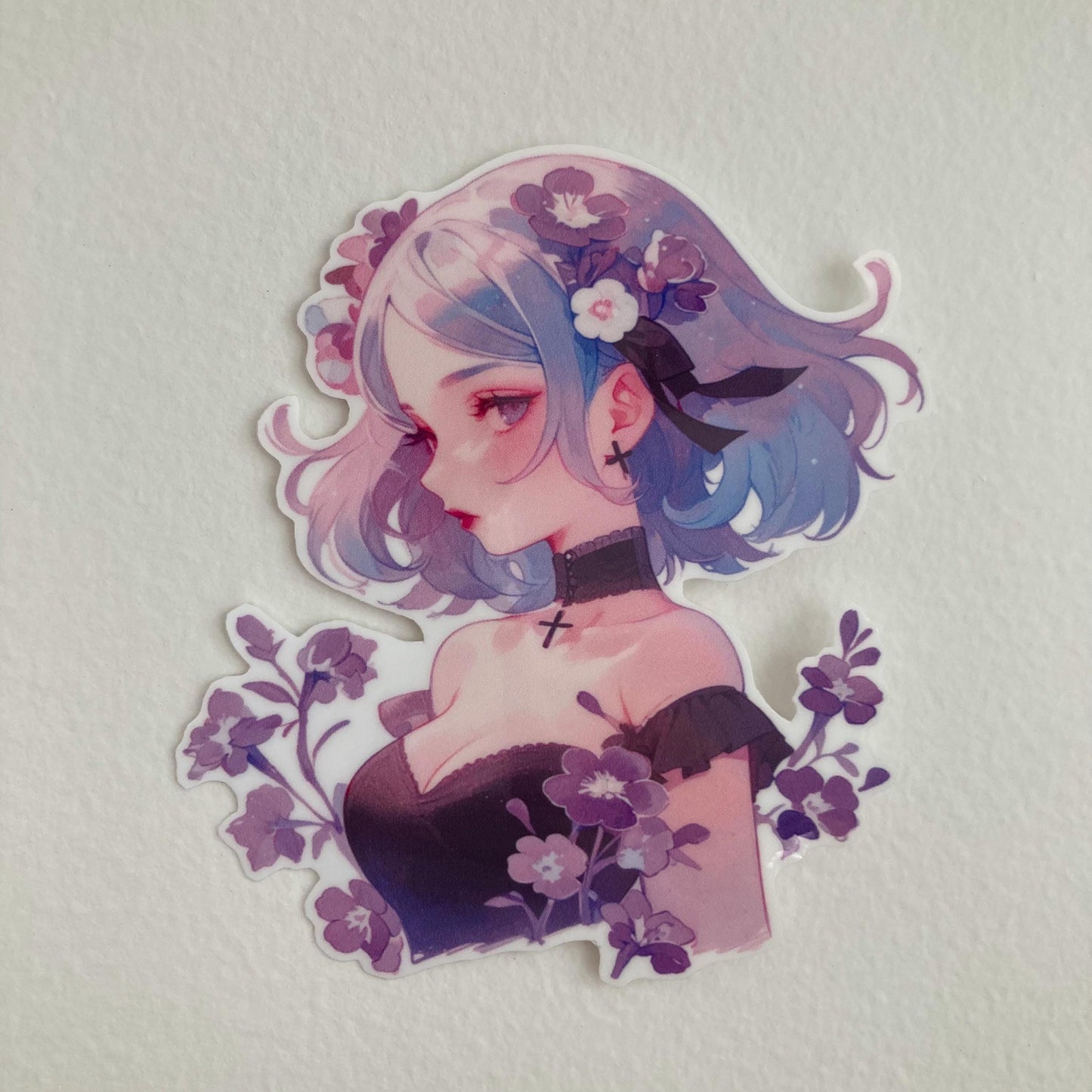 A sticker of a girl with blue and purple hair, wearing a black choker and surrounded by purple flowers, placed on a plain light-colored surface.