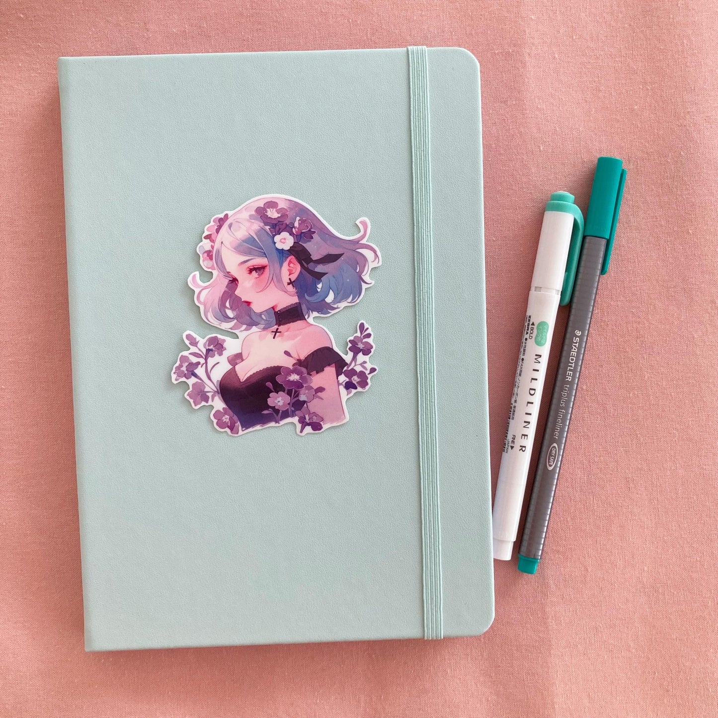 A sticker of a girl with blue and purple hair, surrounded by flowers, is placed on a light teal notebook. Beside the notebook are a green marker and a gray pen, all on a soft pink fabric background.
