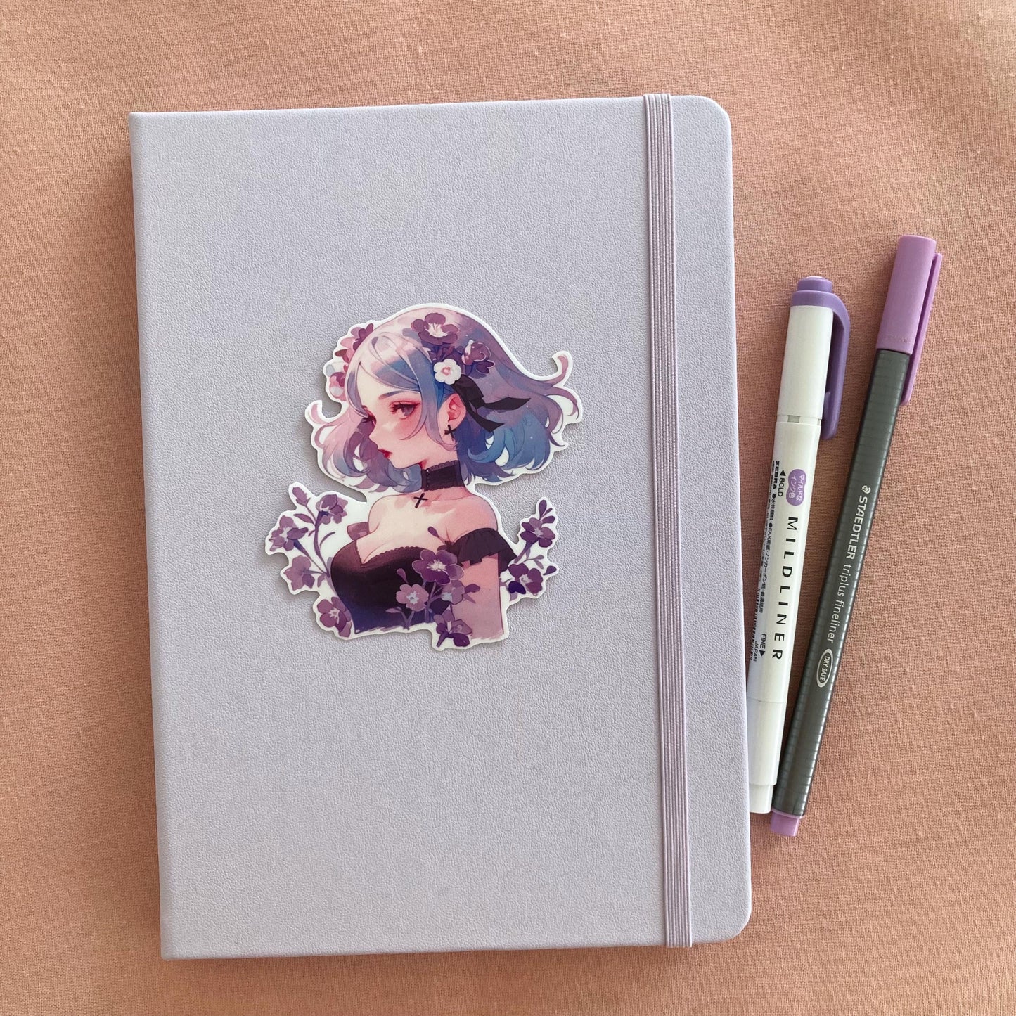 A sticker of a girl with blue and purple hair, surrounded by purple flowers, is placed on a light lavender notebook. A purple marker and gray pen are beside the notebook on a soft pink fabric background.