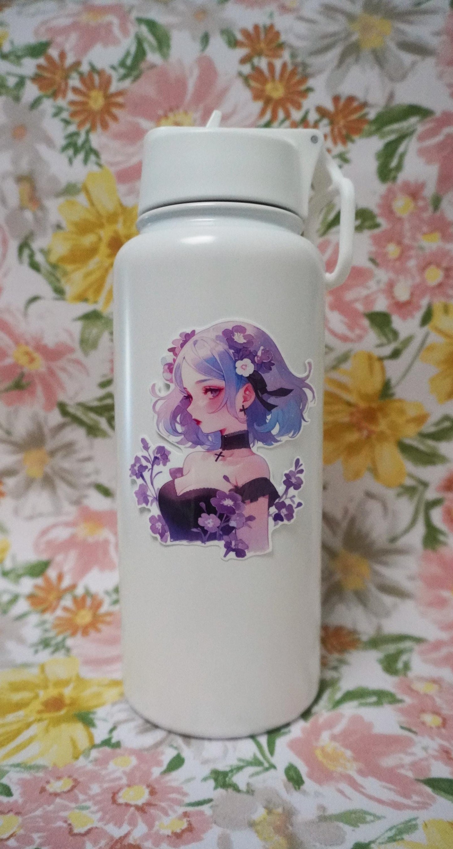 A white water bottle decorated with a sticker of a girl with blue and purple hair, wearing a choker and surrounded by purple flowers. The background is a floral fabric with large pink, yellow, and orange flowers and green leaves.