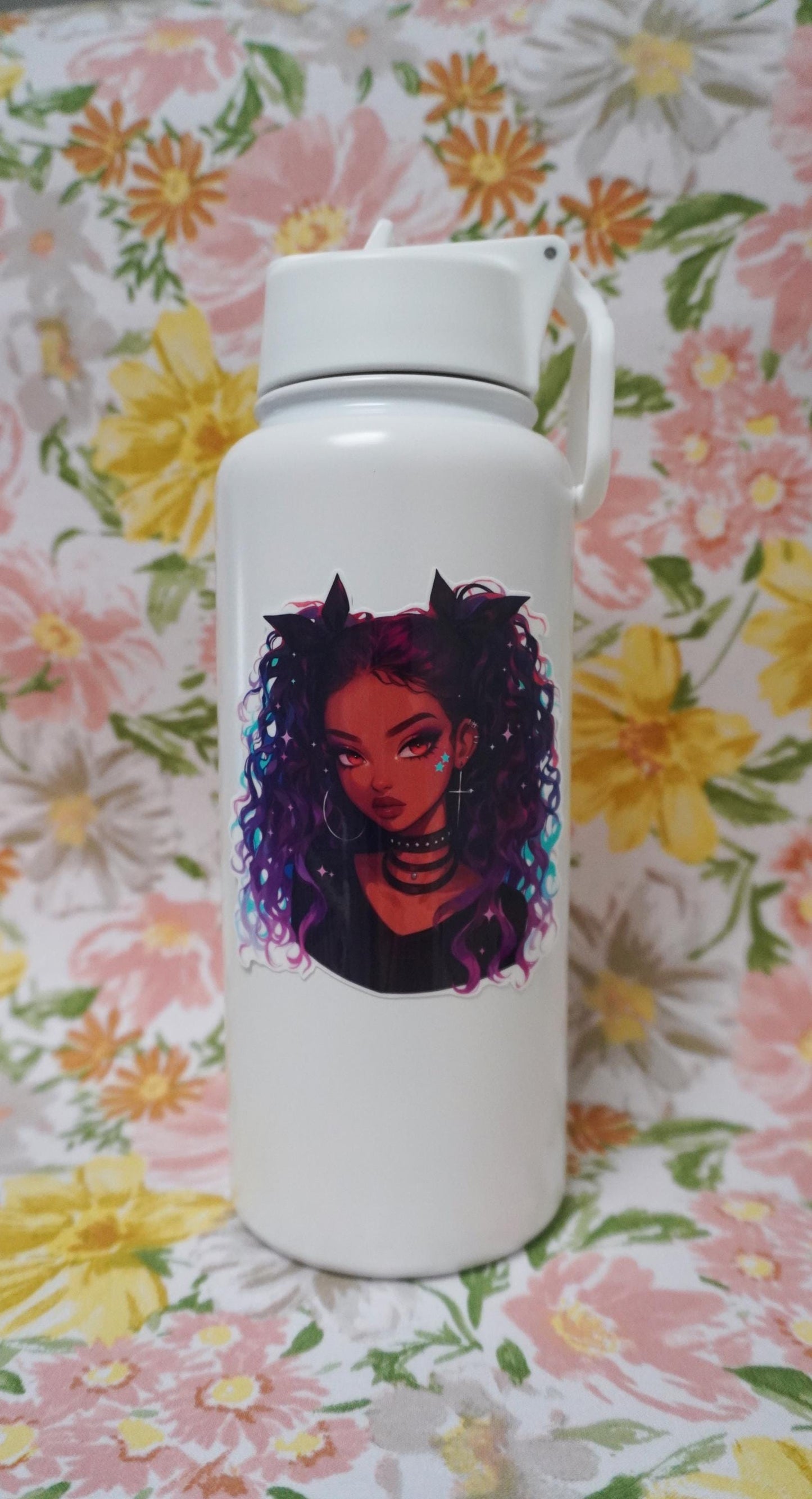 A white water bottle with a sticker of a girl with curly purple and blue hair, chokers, and star face decals. The background is a floral fabric with large pink, yellow, and orange flowers, and green leaves.