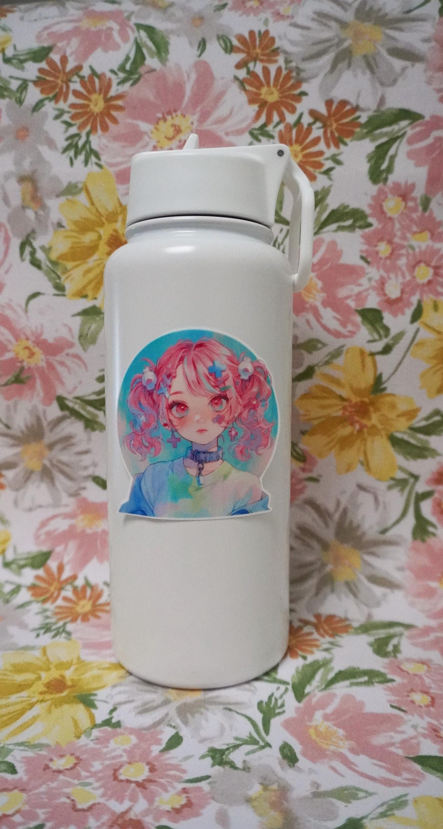 A white water bottle with a sticker of a girl with pink pigtails, wearing a collar and pastel clothes. The background is a floral fabric with pink, yellow, and orange flowers and green leaves.