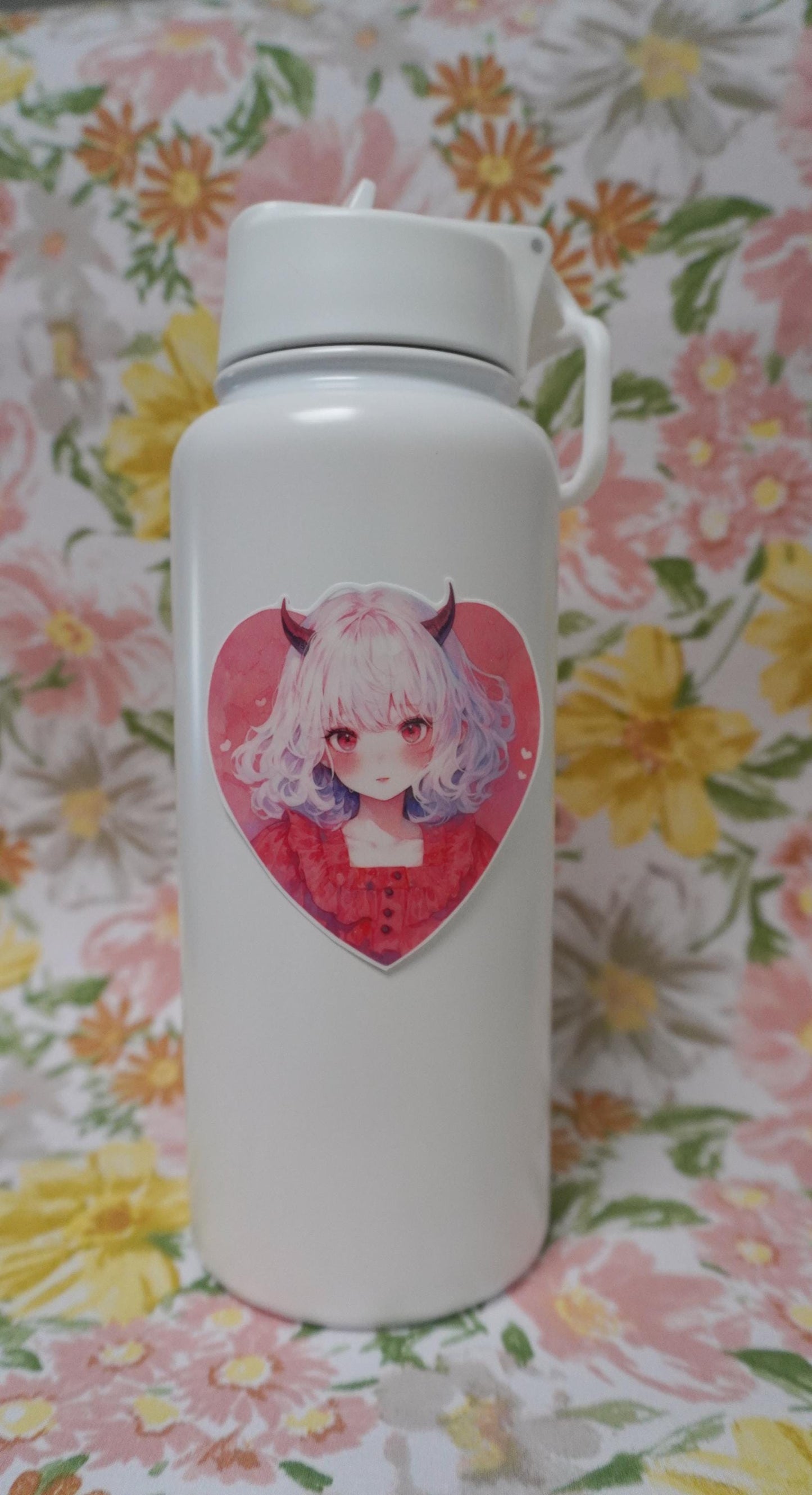A white water bottle with a heart-shaped sticker of a girl with white hair, red horns, and a red dress. The background is a floral fabric with pink, yellow, and orange flowers and green leaves.