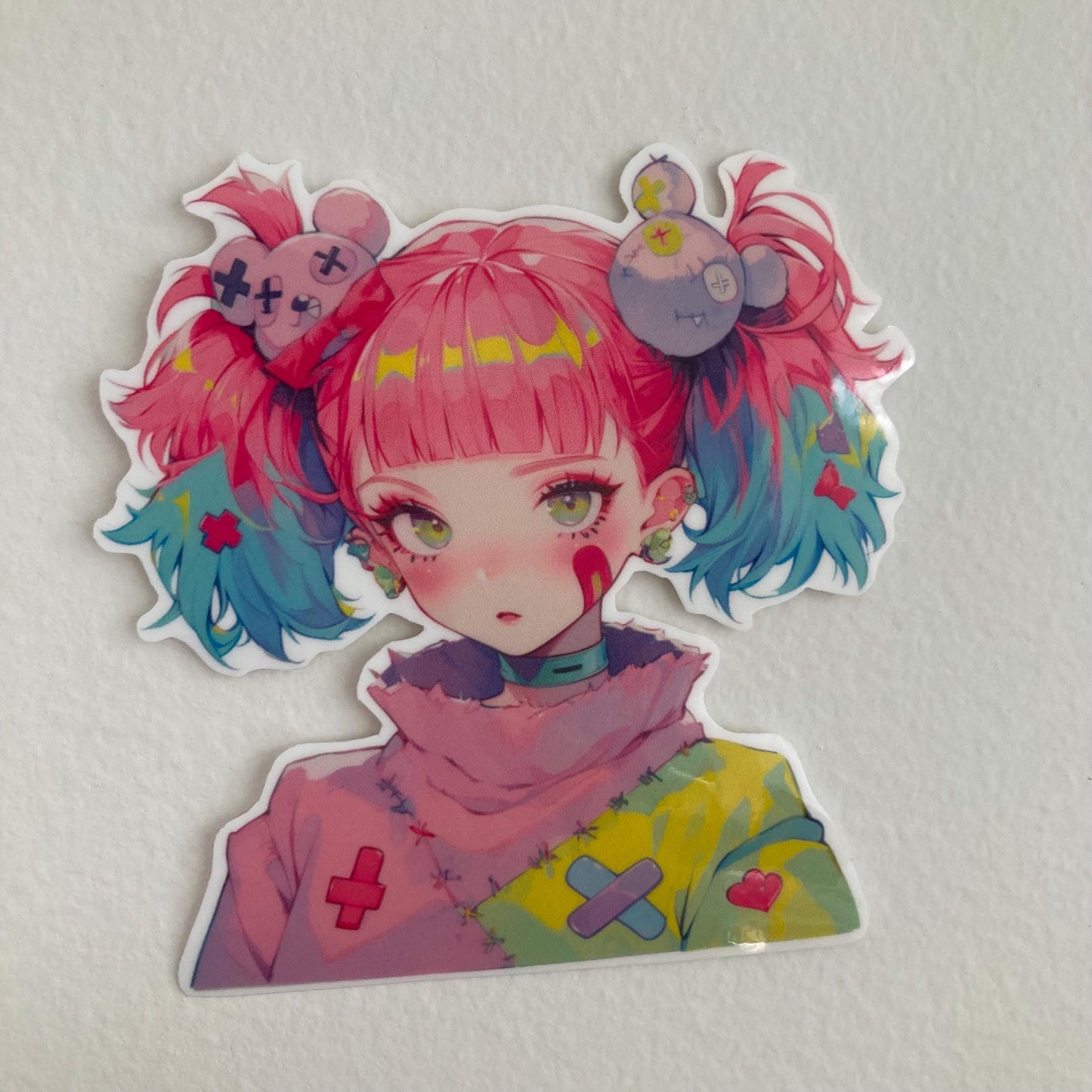 A colorful anime-style sticker of a girl with pink and blue pigtails, wearing a pastel sweater with patches, placed on a plain white surface.