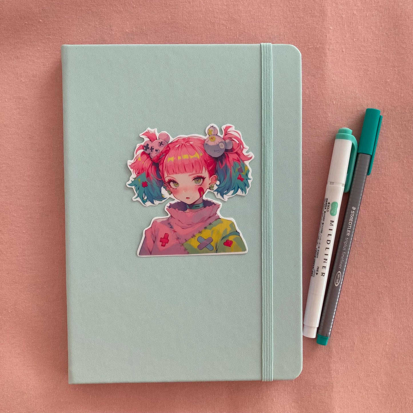 A pastel green notebook with an anime-style sticker of a girl with pink and blue pigtails on its cover, next to a green marker and gray pen, all set on a pink fabric background.