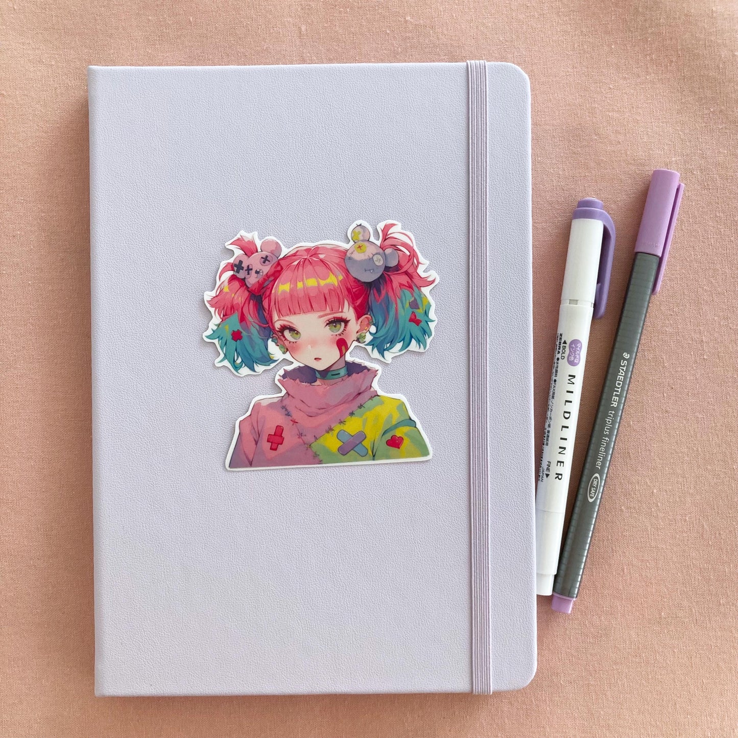 A pastel lavender notebook with an anime-style sticker of a girl with pink and blue pigtails on its cover, next to a purple marker and gray pen, set on a pink fabric background.