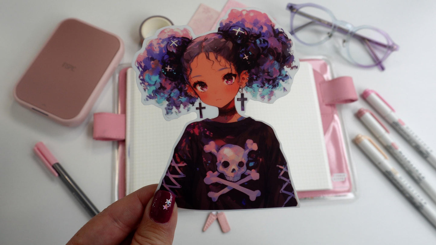 A hand holding a sticker of an anime-style character with purple and blue curly hair in pigtails, wearing cross earrings and a skull-themed shirt. The background includes pink stationery, an open notebook, and pastel markers on a white surface.