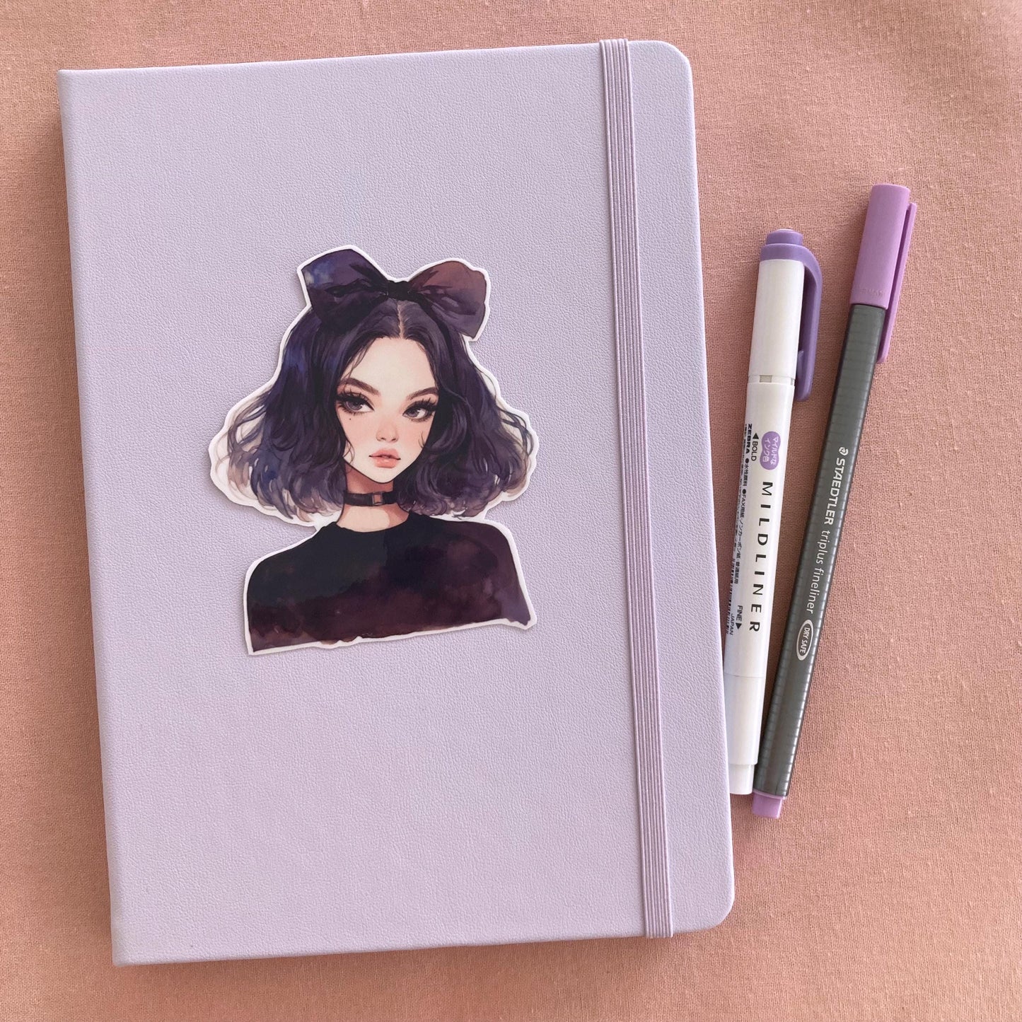 A pastel lavender notebook with a sticker of an anime-style character with dark purple hair, a large bow, and a black choker, next to a purple marker and gray pen on a pink fabric background.