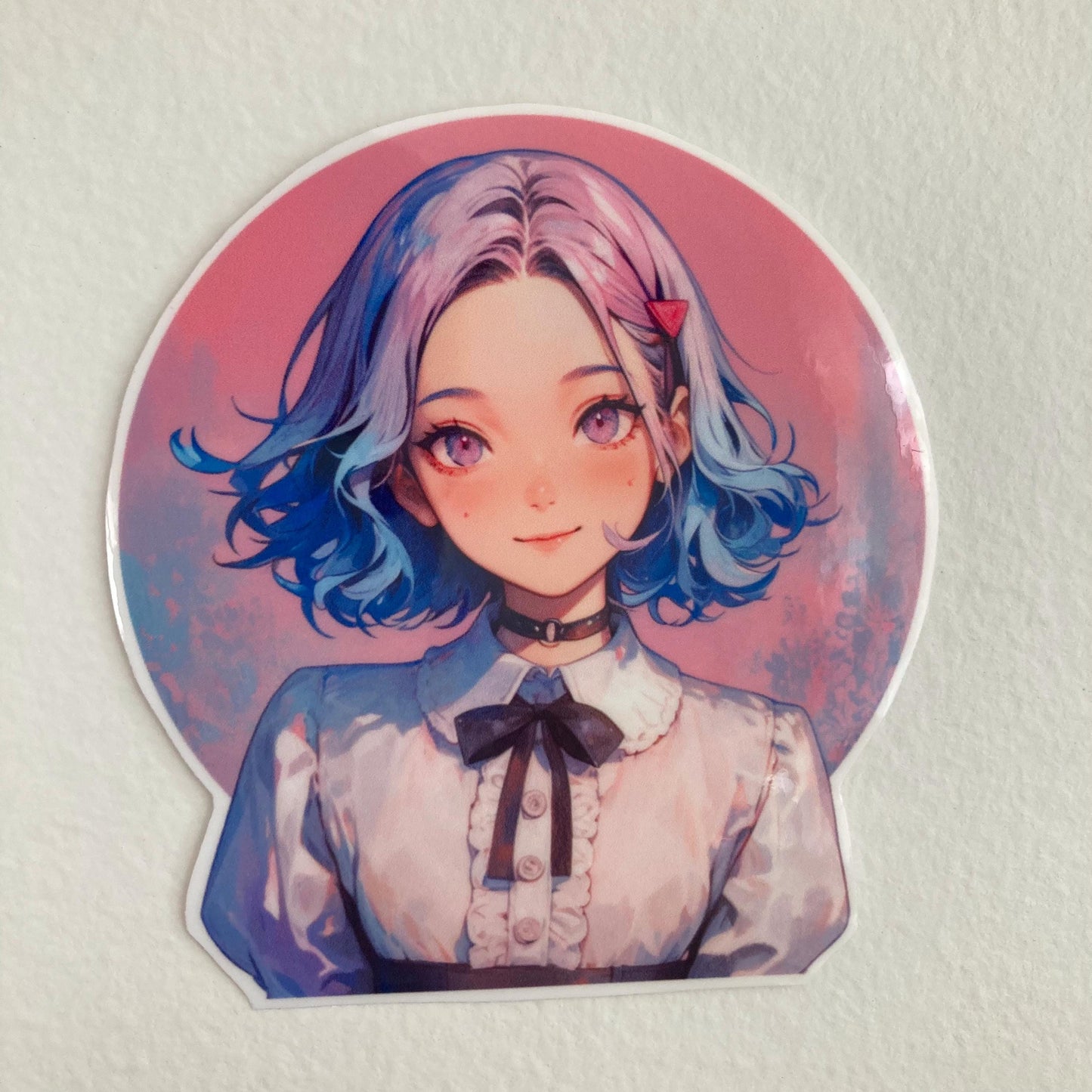 A round sticker featuring an anime-style character with short, wavy blue and pink hair, pink eyes, and a hair clip. The character wears a white blouse with ruffles and a black bow, against a pink gradient background.
