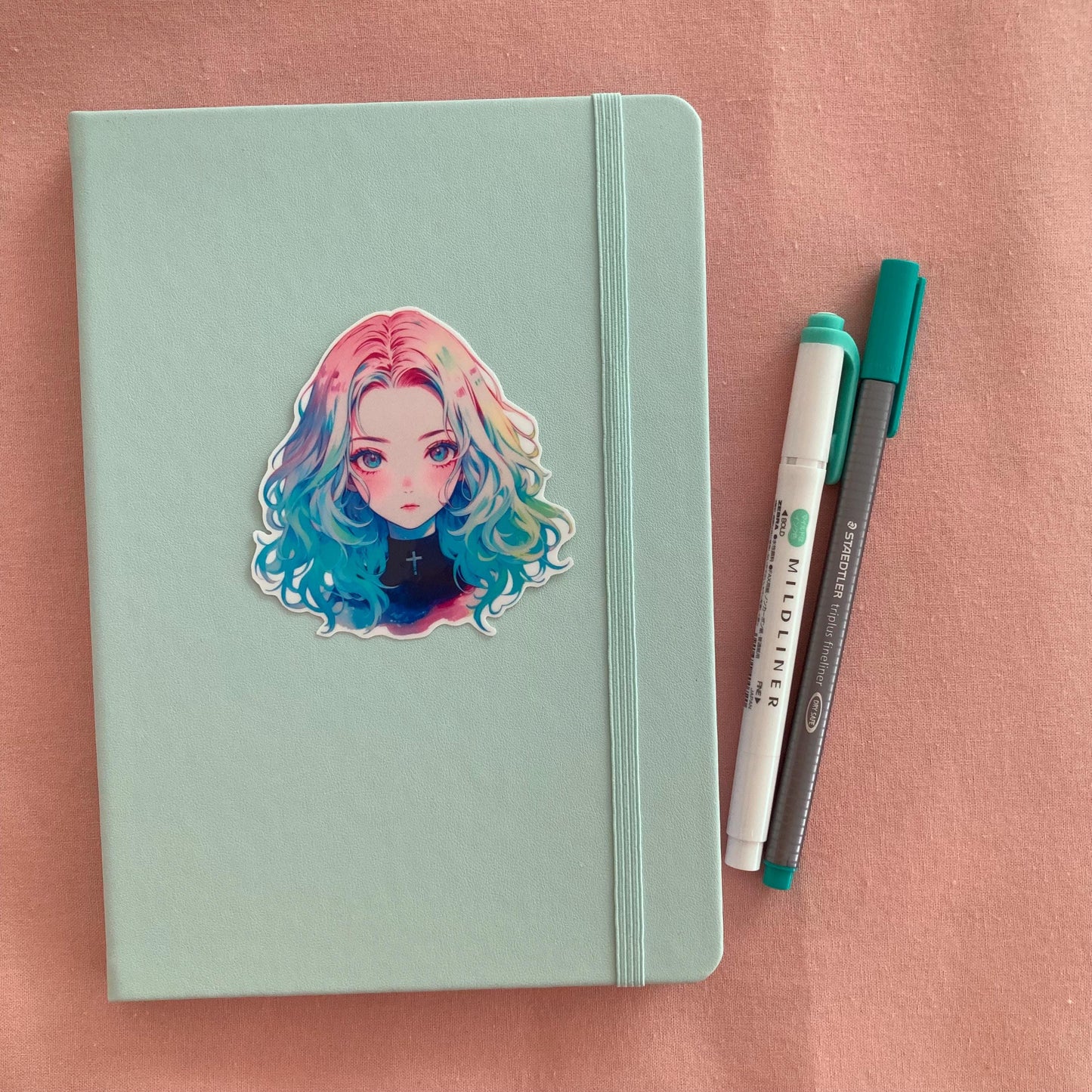 Mint green notebook with an anime sticker of a girl with rainbow hair, placed on a pink surface. A teal pen and a white highlighter lie beside the notebook.