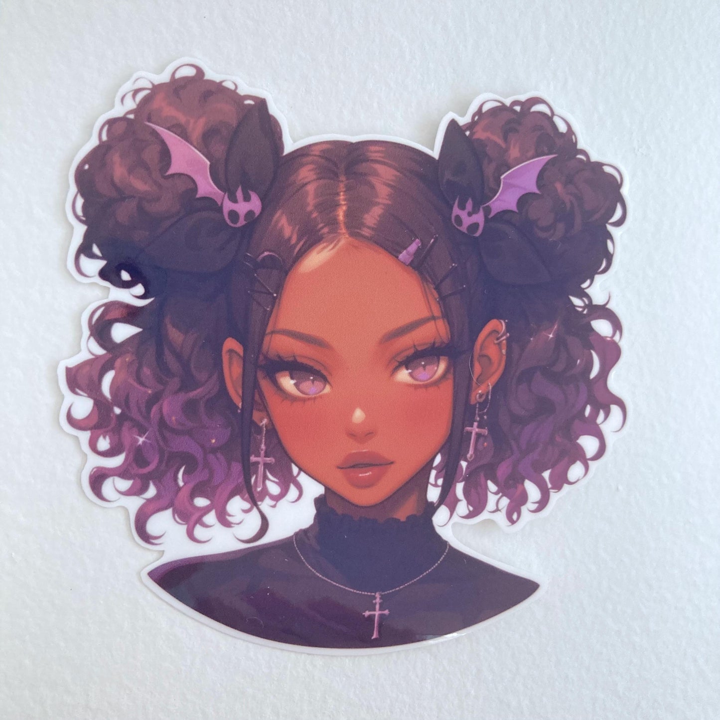 A sticker of an illustrated girl with curly hair in buns adorned with bat-shaped hair clips. She has purple eyes, cross earrings, and is wearing a black top. The background is a plain, textured white surface.