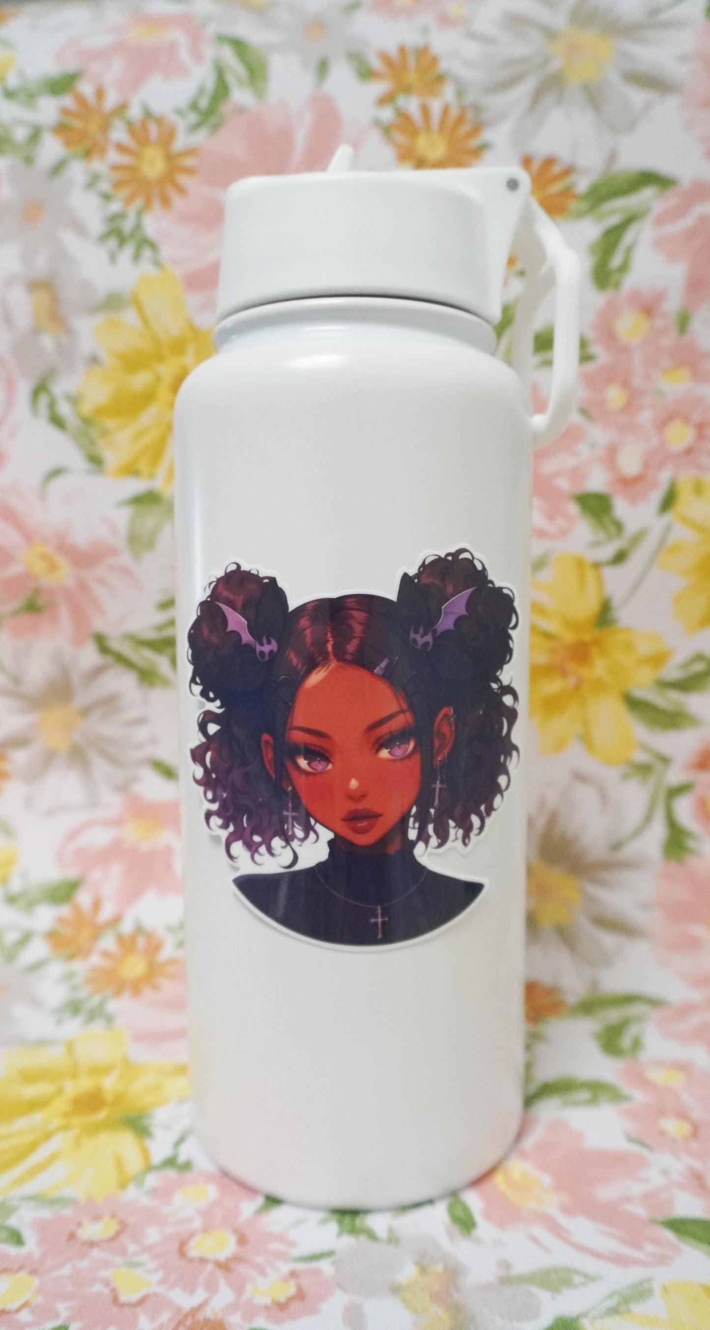 A white bottle with a sticker of an illustrated girl with dark curly hair styled in buns, wearing bat-shaped hair clips and cross earrings. The background is a floral pattern with large yellow, pink, and orange flowers and green leaves.