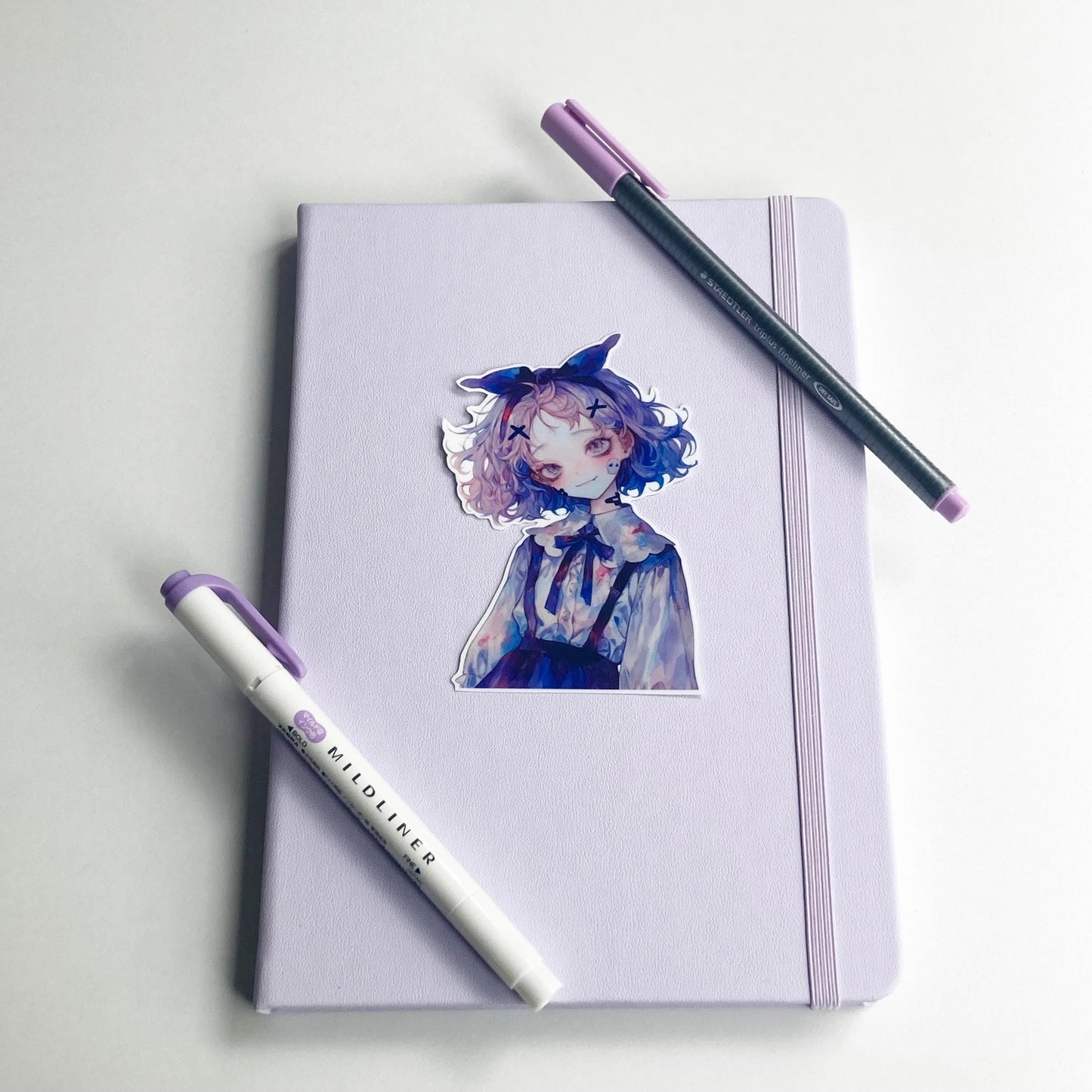 A sticker of an anime-style character is placed on a closed lavender notebook with an elastic band. Two pens, one gray with a purple cap and one white "Mildliner" marker with a purple cap, are laid diagonally across the notebook.