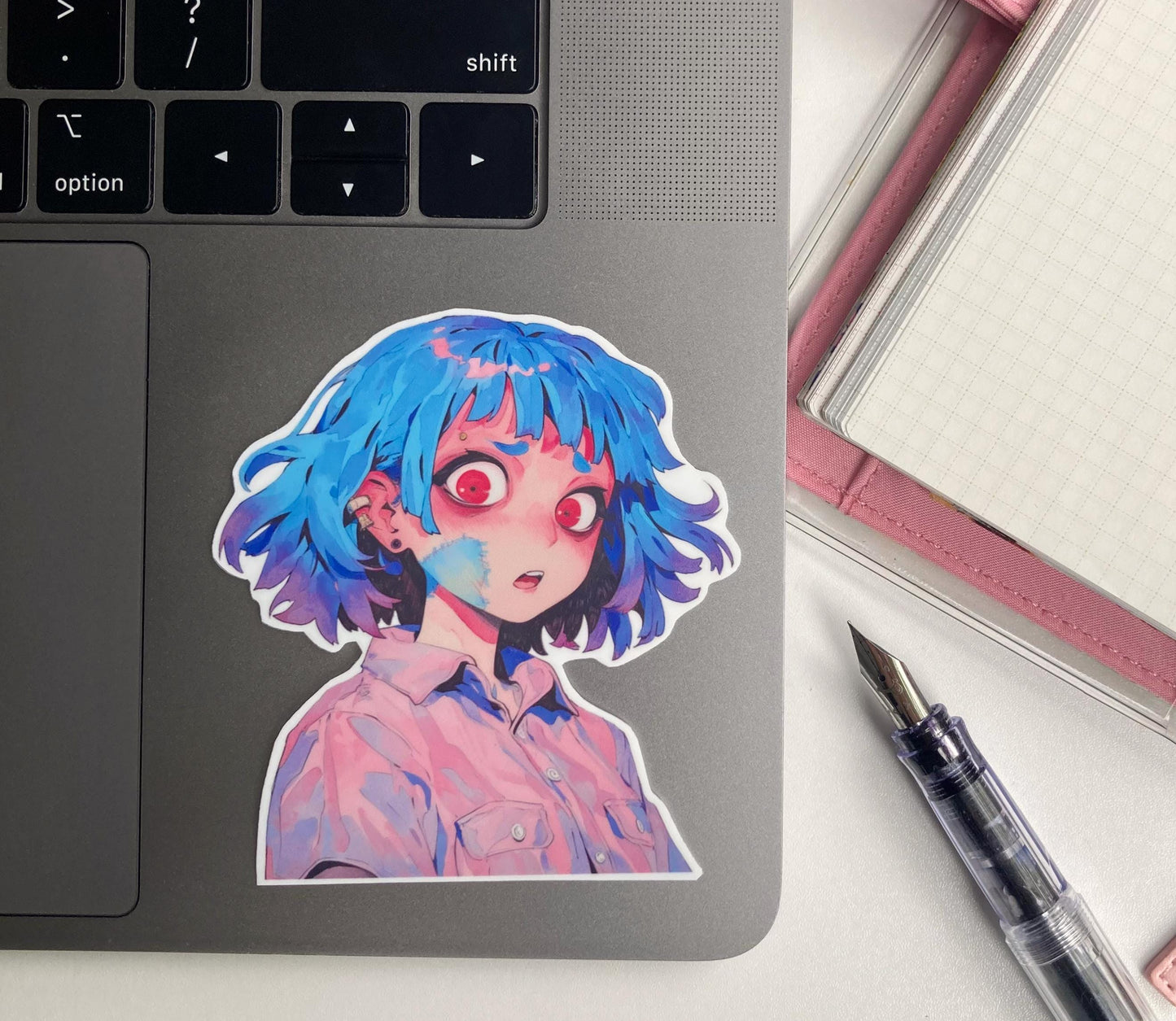 A sticker of an anime character with blue hair and red eyes is placed on the surface of a laptop keyboard. To the right, a pink notebook and a transparent fountain pen are partially visible. The keyboard's "option" and arrow keys can be seen.