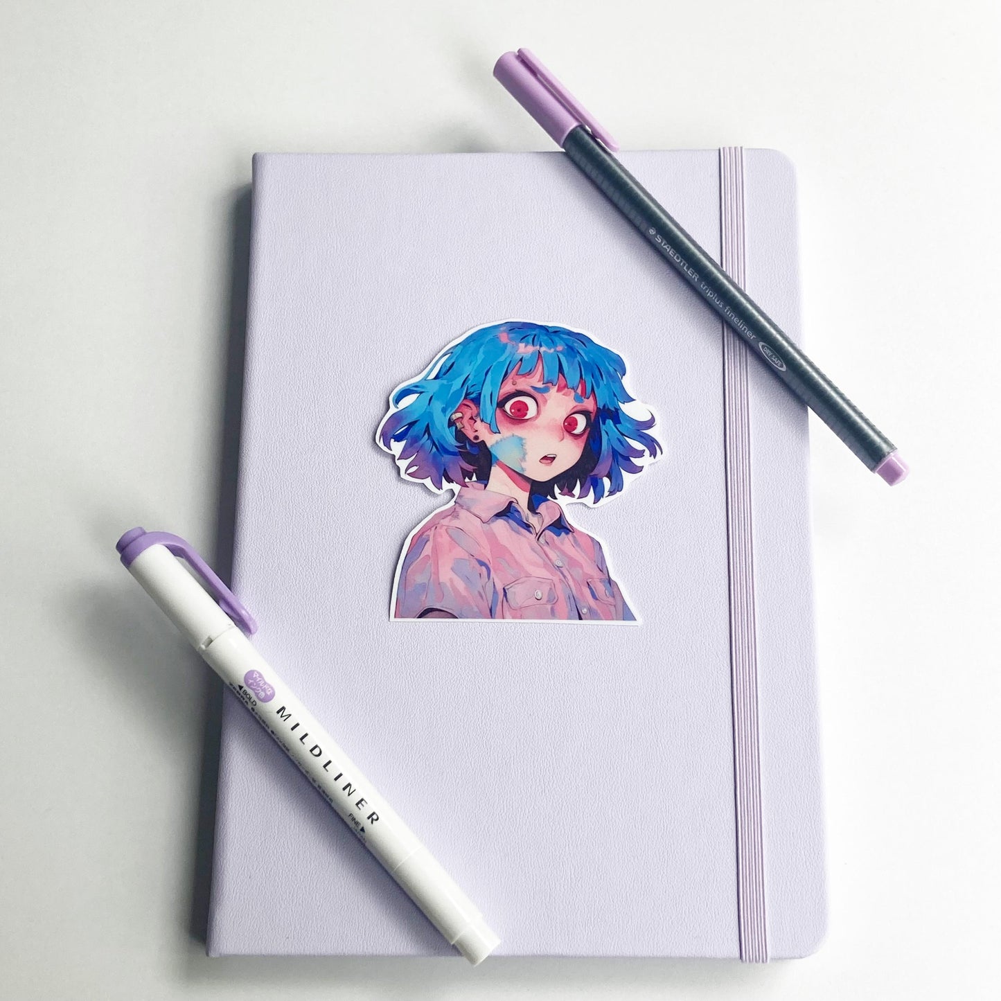 A sticker of an anime character with blue hair and red eyes is placed on a closed light purple notebook. A purple marker and a dark gray pen with a purple cap lie diagonally across the notebook. The background is a white surface.