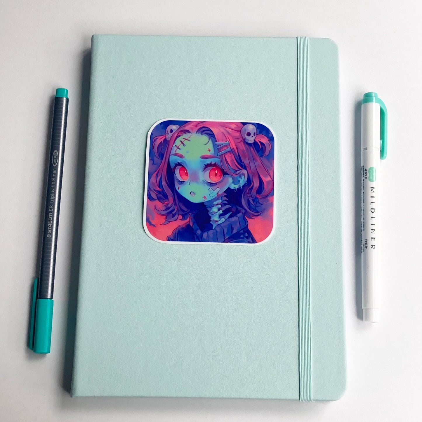A square sticker of an anime character with pink hair, red eyes, and skull accessories is placed on a closed light teal notebook. A dark gray pen with a teal cap is on the left, and a teal marker is on the right. The background is a white surface.