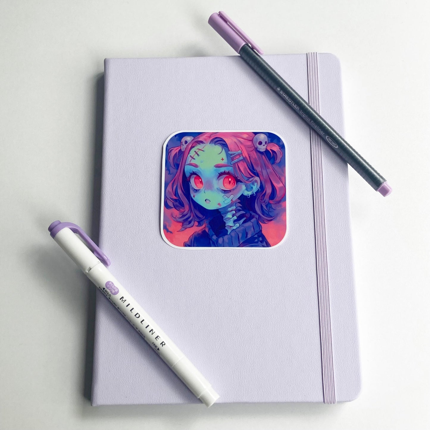 A square sticker of an anime character with pink hair, red eyes, and skull accessories is placed on a closed light purple notebook. A purple marker and a dark gray pen with a purple cap lie diagonally across the notebook.