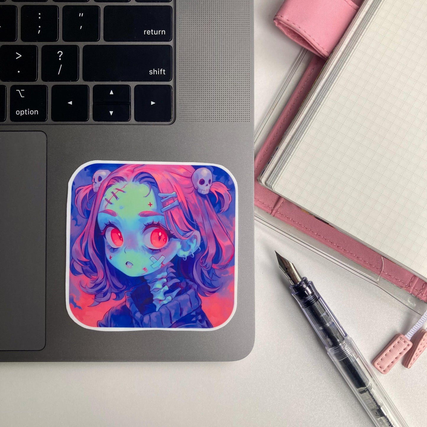 A square sticker of an anime character with pink hair, red eyes, and skull accessories is placed on a laptop keyboard. To the right, a pink notebook and a transparent fountain pen are visible. The keyboard's "shift" and arrow keys are shown.