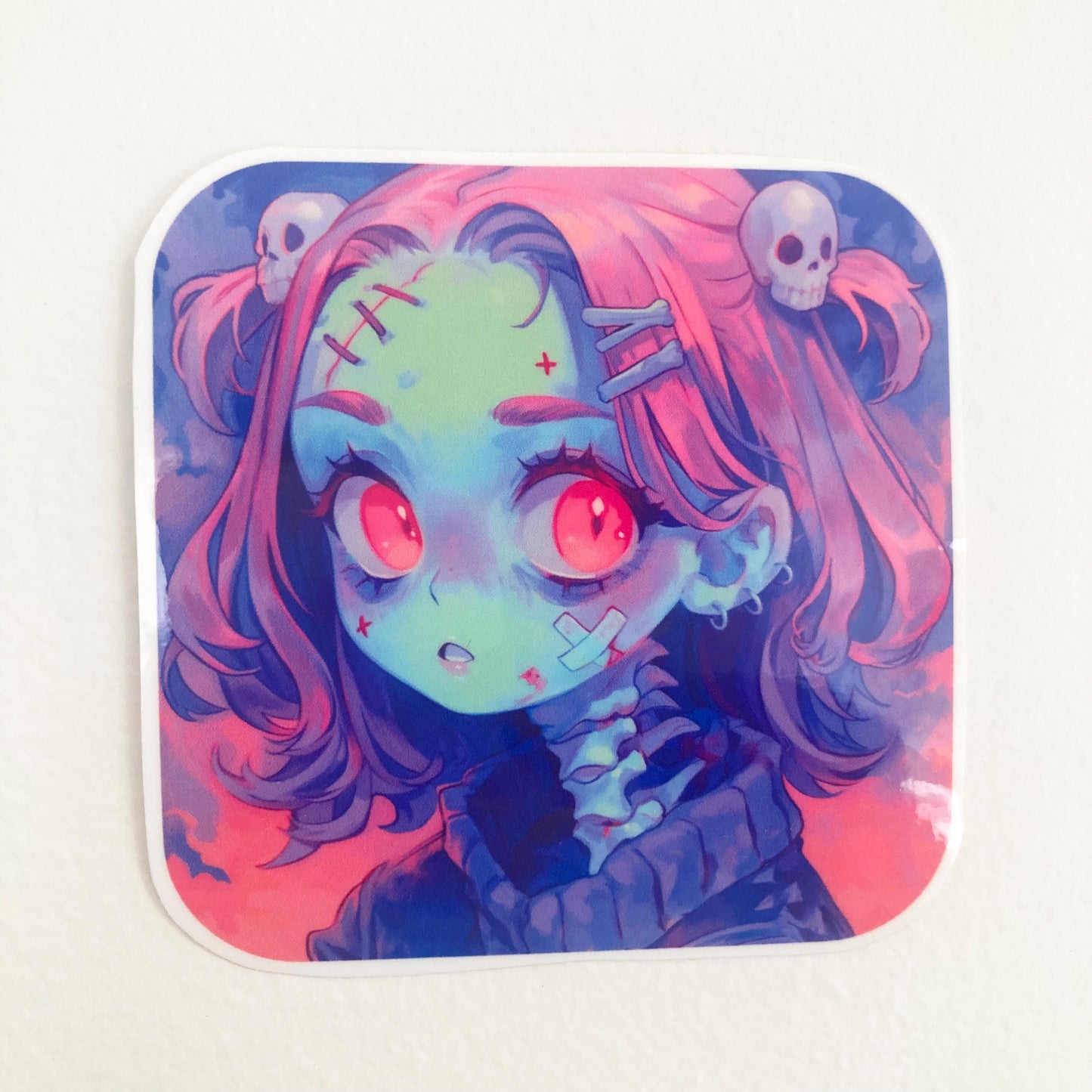 A close-up of a square sticker featuring an anime character with pink hair, red eyes, and a greenish face, adorned with skull accessories and stitched details. The sticker is placed on a plain, light-colored background.