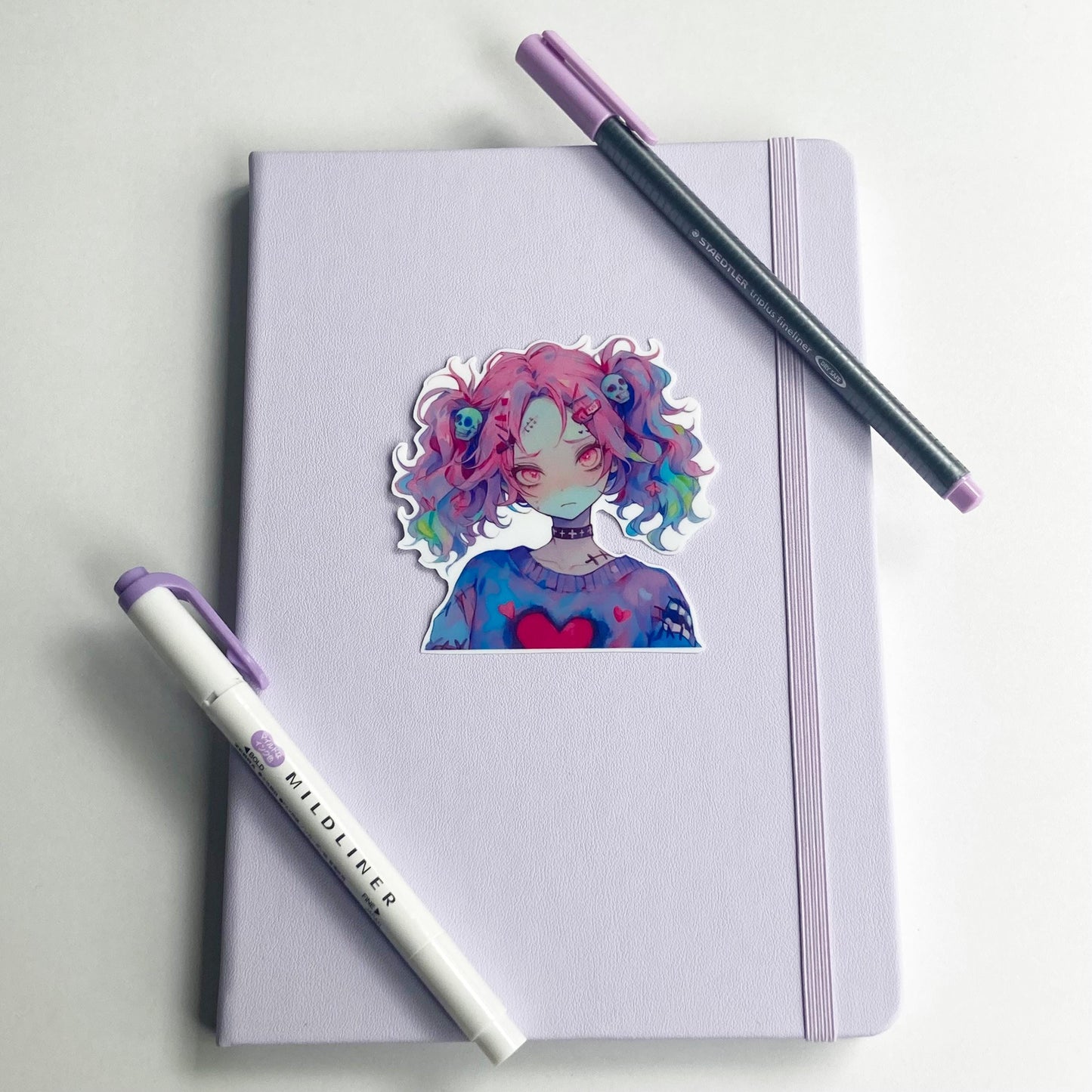 A sticker of an anime character with pink and multicolored hair and skull accessories is placed on a light purple notebook. A purple marker and a pen with a purple cap are positioned diagonally on the notebook.