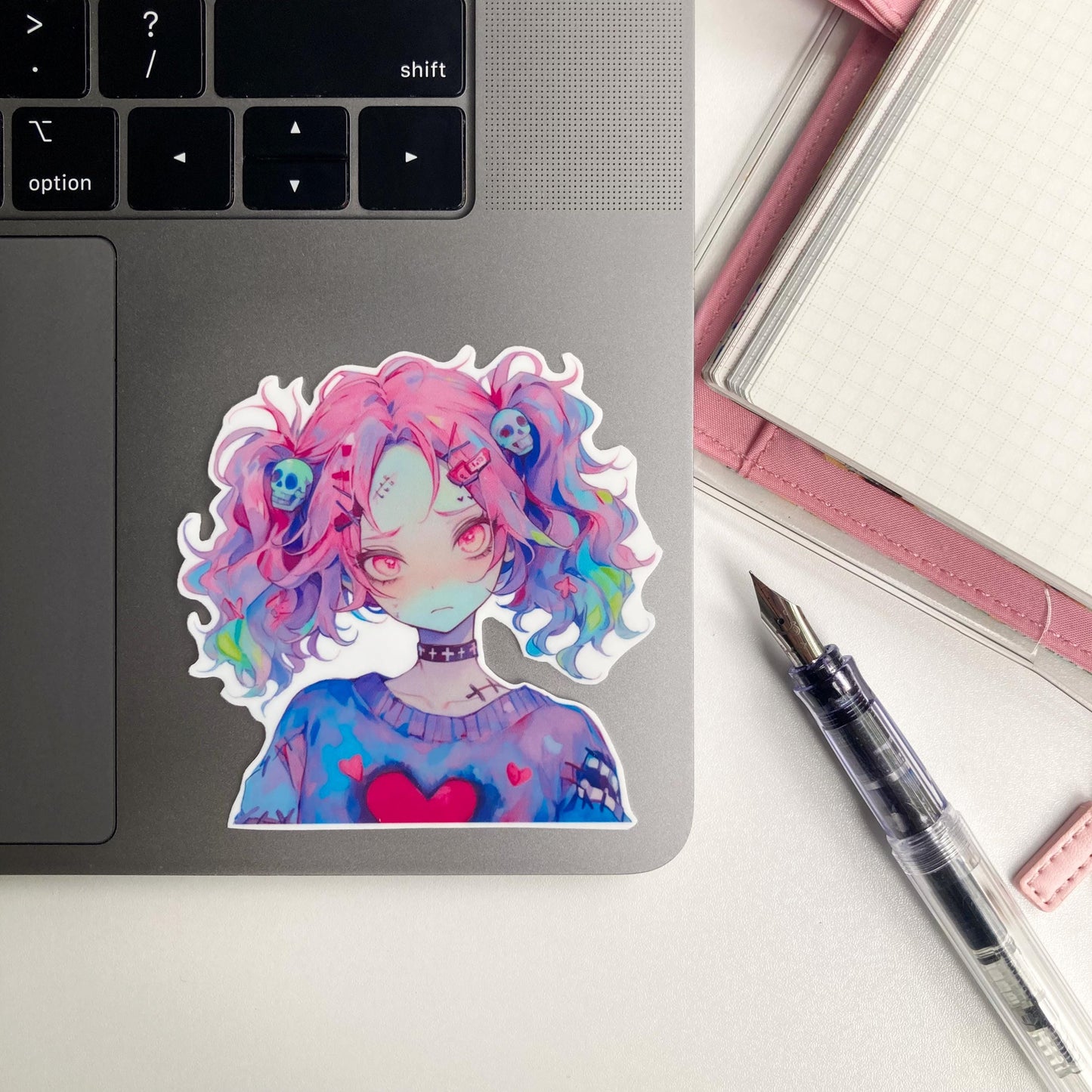 A sticker of an anime character with pink and multicolored hair, skull accessories, and a heart on their sweater is placed on a laptop. A pink notebook and a fountain pen are nearby.