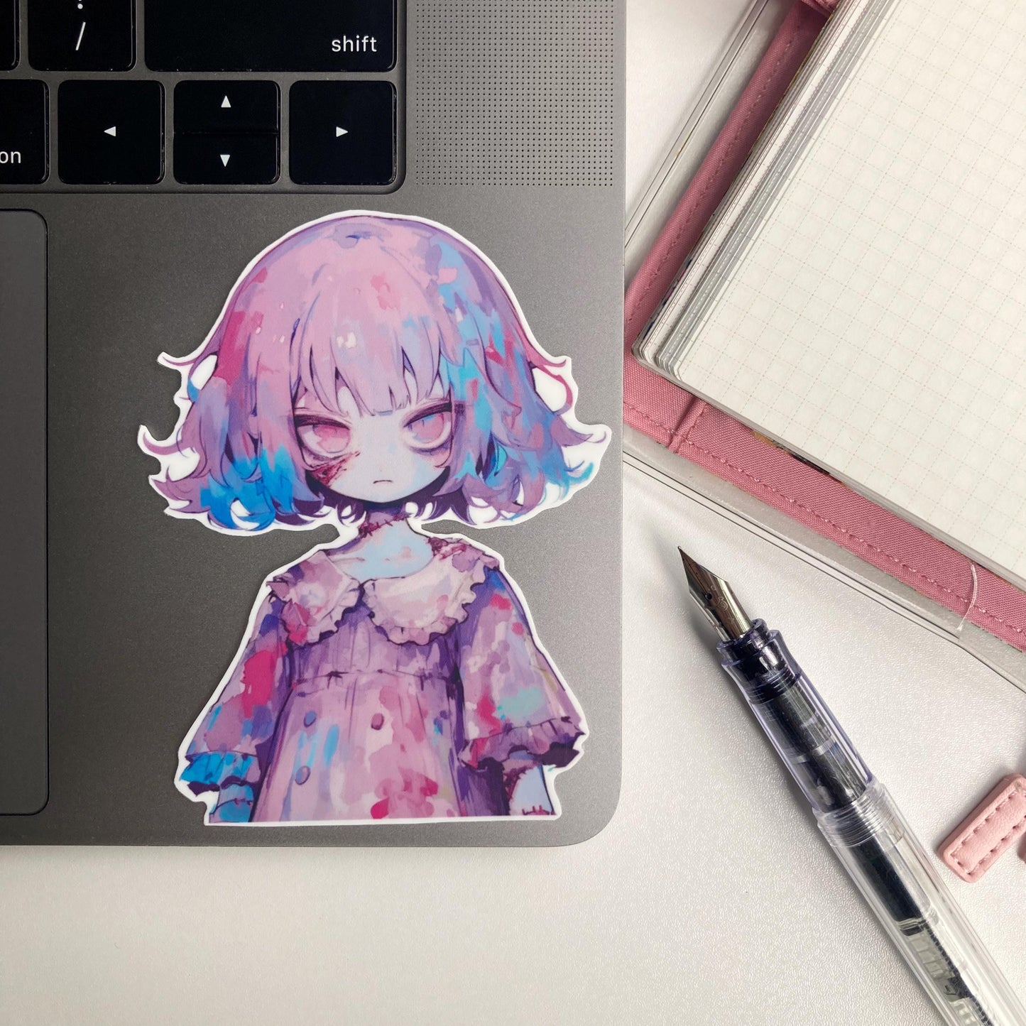 A sticker of an anime character with purple and blue hair, wearing a ruffled dress, is placed on a laptop. A pink notebook and a fountain pen are nearby.