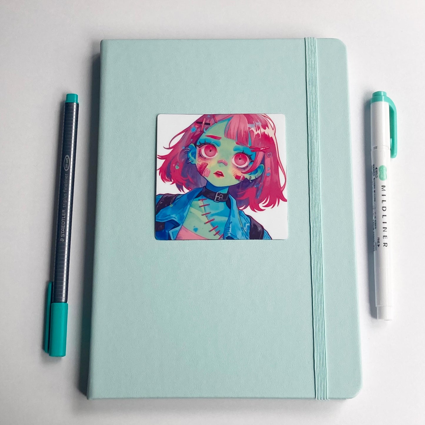 A mint green notebook with a sticker of an illustrated character with pink hair, green skin, and stitched details. A teal-capped gray pen is on the left, and a teal-tipped Mildliner marker is on the right, both placed parallel to the notebook
