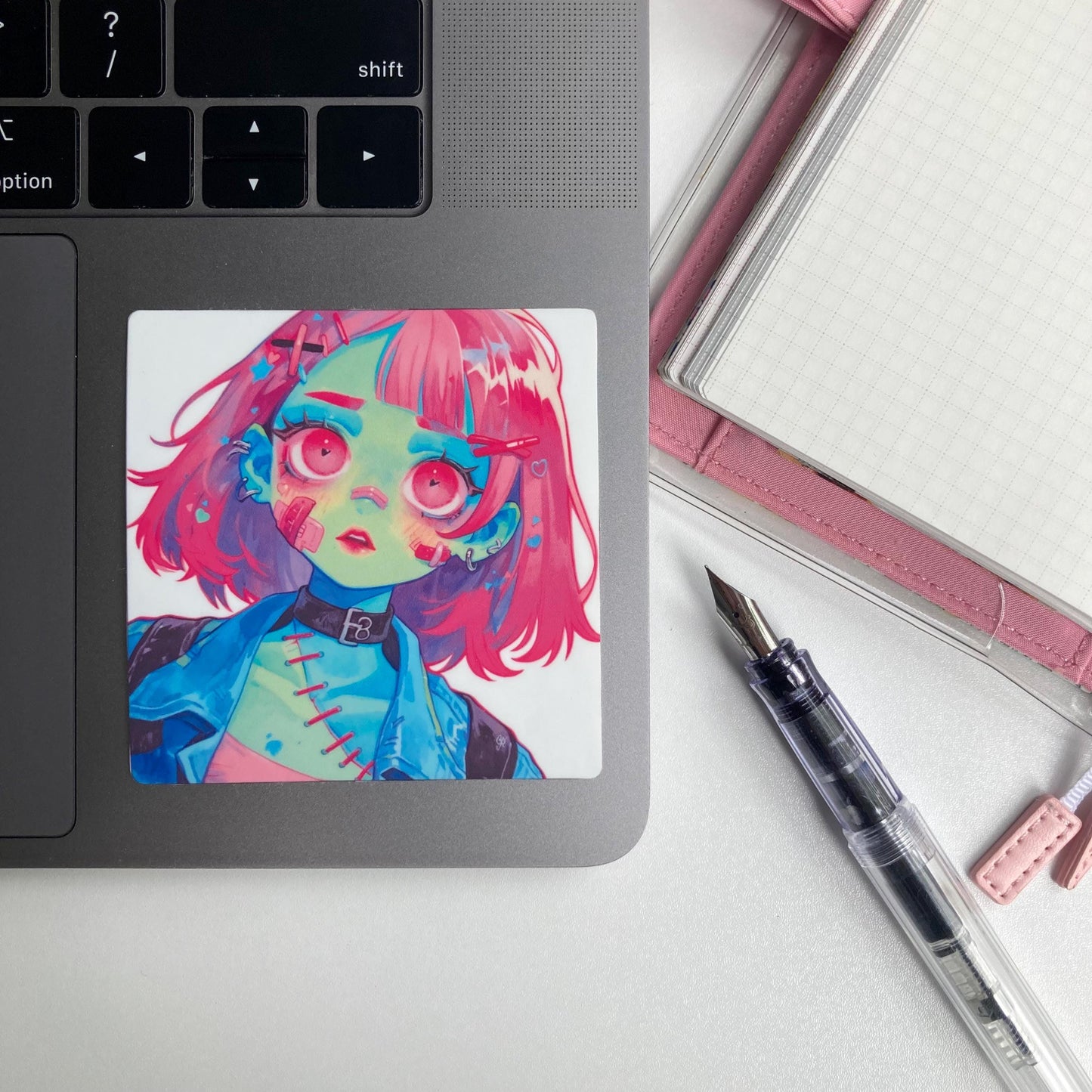 A square sticker of an anime character with pink hair, green skin, and large pink eyes is placed on a laptop. Nearby are a pink notebook and a fountain pen.