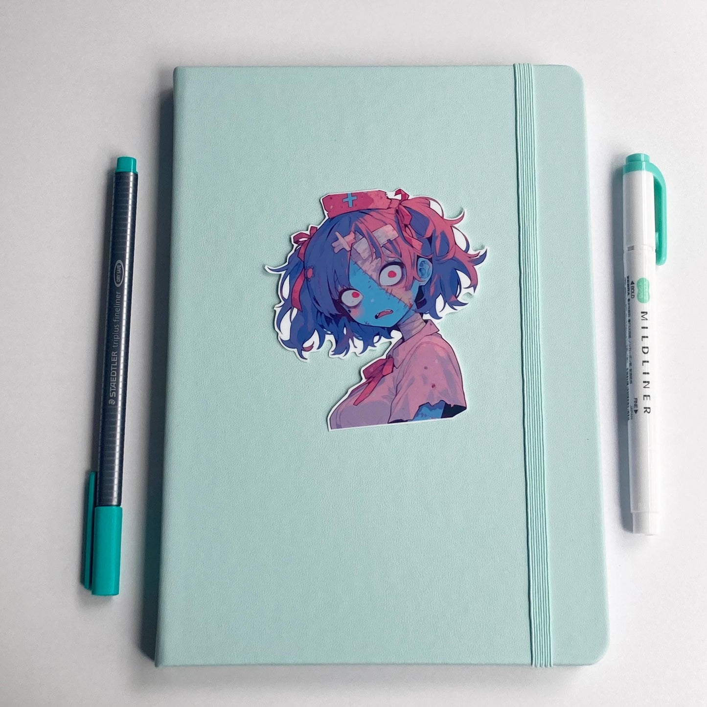 A blue and pink skinned zombie nurse sticker is on a mint notebook. The notebook has a highlighter and pen sitting next to it. The sticker has purple and pink pigtails in bows. Wears a pink nurses outfit with tears and a nurse hat with cross