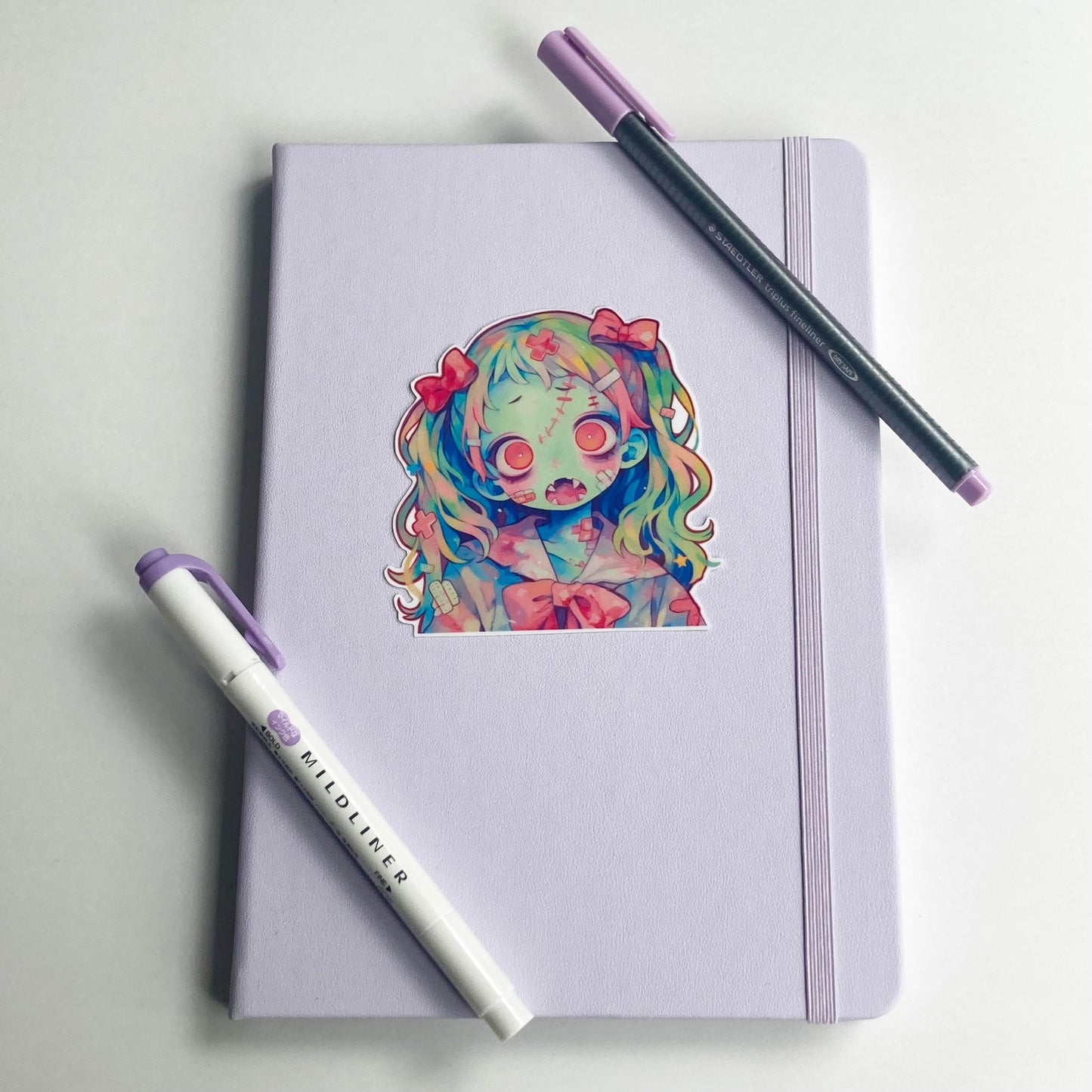 Sticker of an anime-style zombie girl with rainbow hair, pink bows, greenish-blue skin, and pink eyes on a lavender notebook. The character has stitches, bandages, and a pink bow on her outfit. A purple pen and highlighter are placed on the notebook.