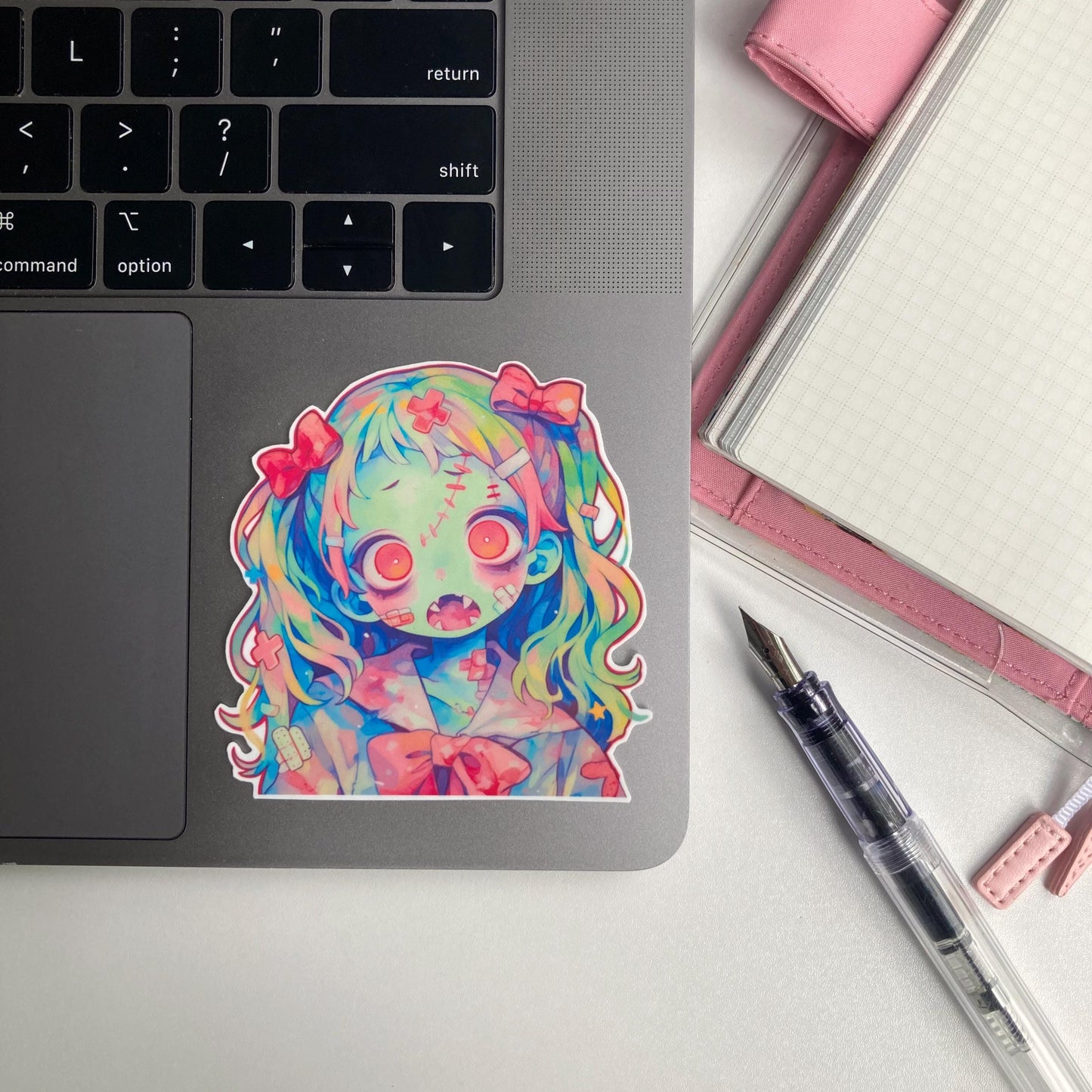 Sticker of an anime-style zombie girl with multicolored hair, pink bows, greenish-blue skin, and pink eyes, featuring stitches and bandages. The character has an open mouth with sharp teeth. The sticker is placed on a laptop keyboard corner.
