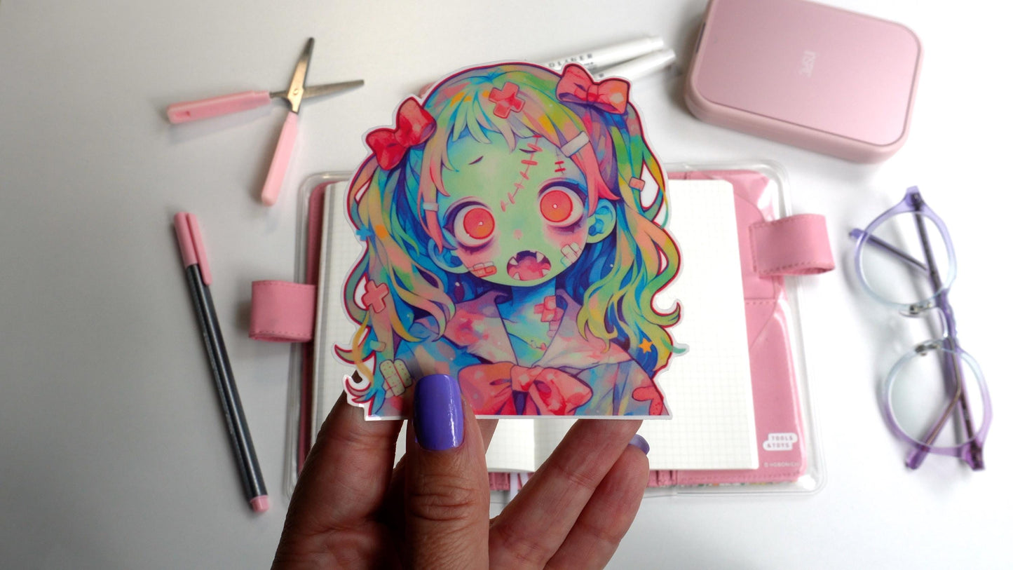 A sticker of an anime-style zombie girl with multicolored hair, pink bows, and a greenish-blue skin tone. She has large pink eyes, an open mouth with sharp teeth, and bandages. The sticker is held up by a hand with purple-painted nails.