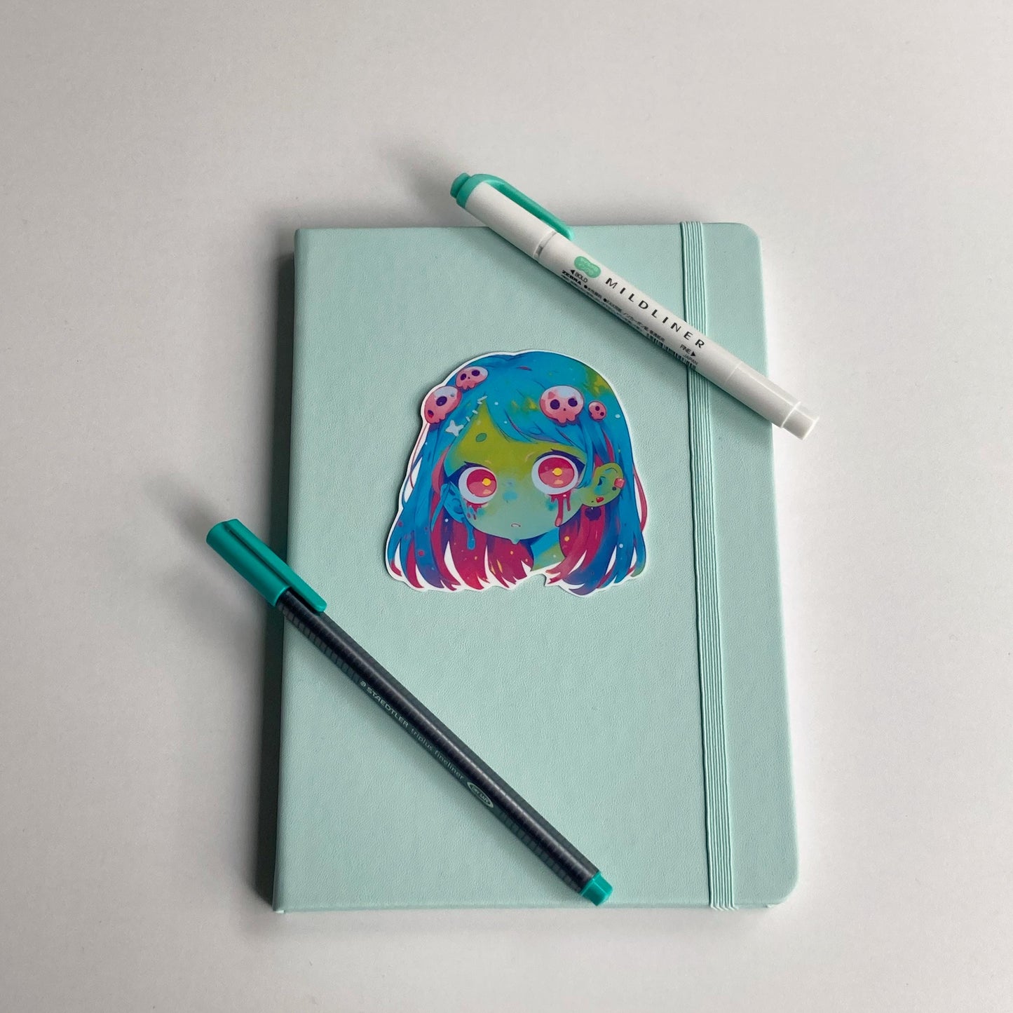 Sticker of an anime zombie girl with blue and pink hair, green skin, and large pink eyes on a light teal notebook. Blood drips from her eyes, and her hair is decorated with small pink skulls. A teal pen and matching highlighter are on the notebook.