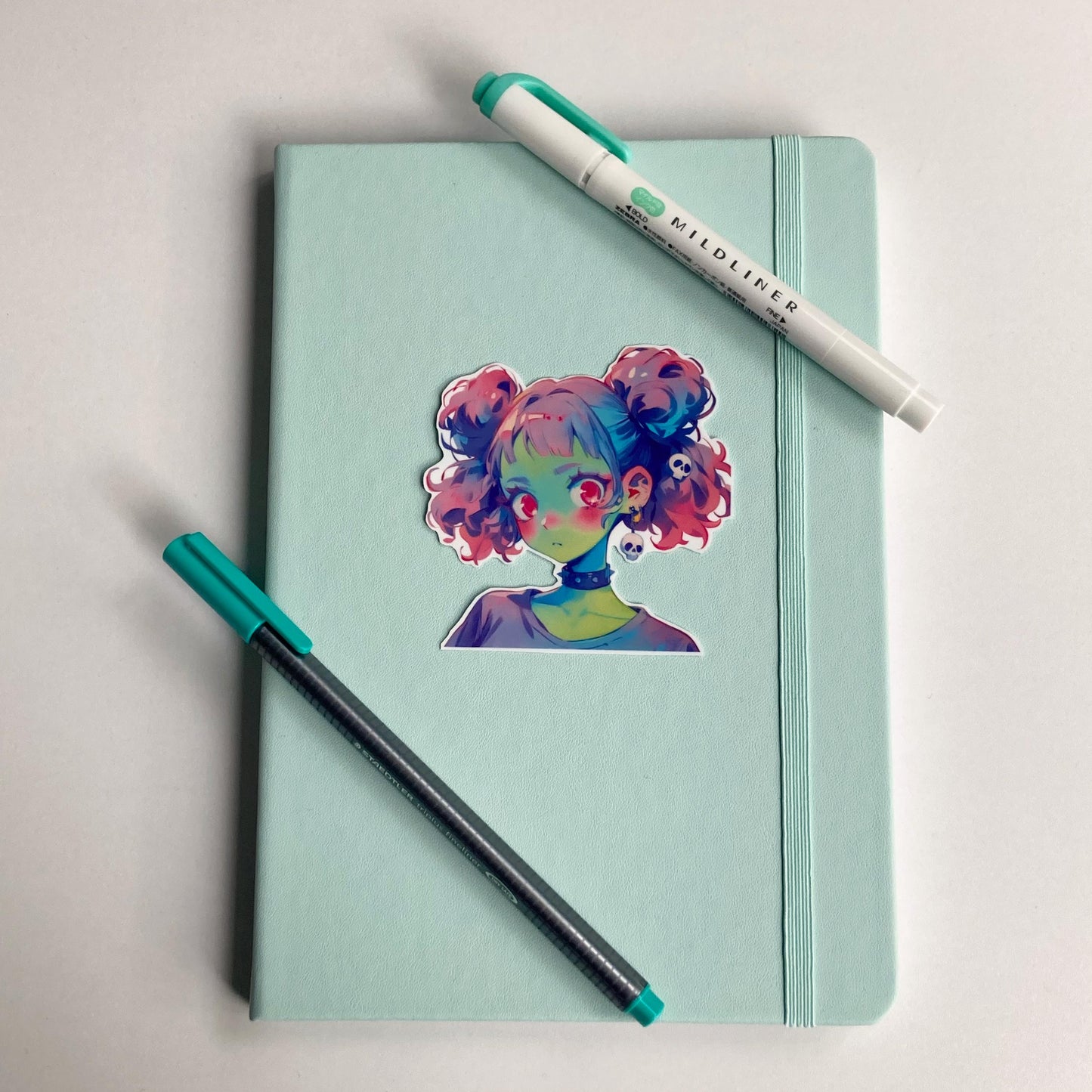 Sticker of an anime-style character with pastel pink and blue twin buns, green skin, and pink eyes on a light teal notebook. The character wears a black choker and skull earrings. A teal pen and matching highlighter are placed on the notebook.