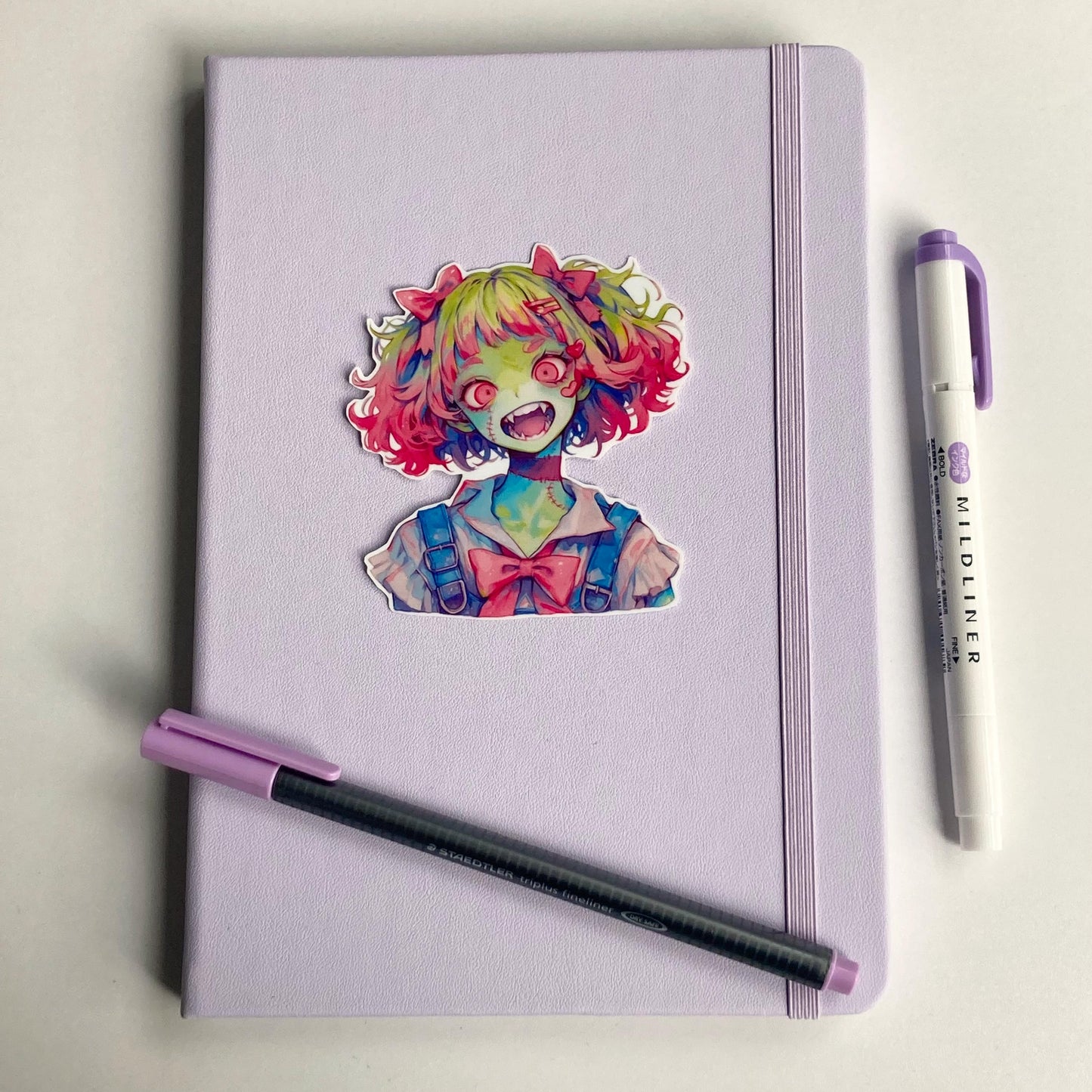 A colorful anime sticker of a zombie girl with green skin, pink and green hair, wide red eyes, and fangs is placed on a lavender notebook.