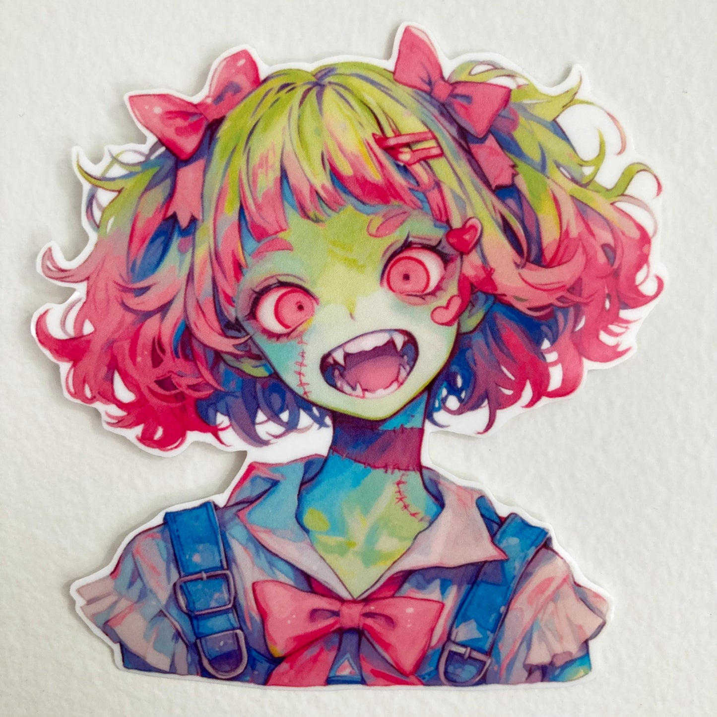 A colorful anime-style sticker of a zombie girl with green skin, wide red eyes, fangs, and pink and green hair. She wears a white collar, blue overalls, and a large pink bow, with her mouth open in an excited expression.