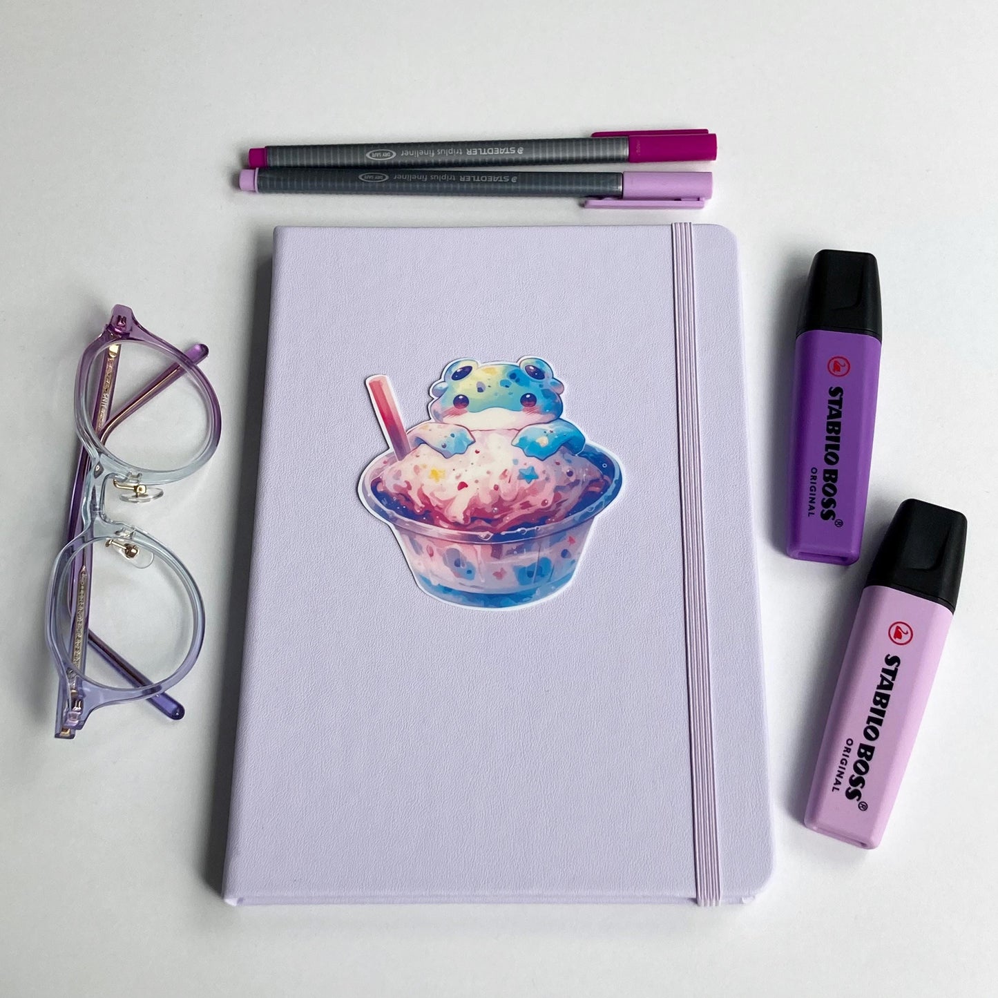 A lavender notebook with a sticker of a colorful alien frog in a cup of pastel ice cream is surrounded by purple-rimmed glasses, two highlighters, and two pens in shades of purple and pink. The items are neatly arranged on a white surface.