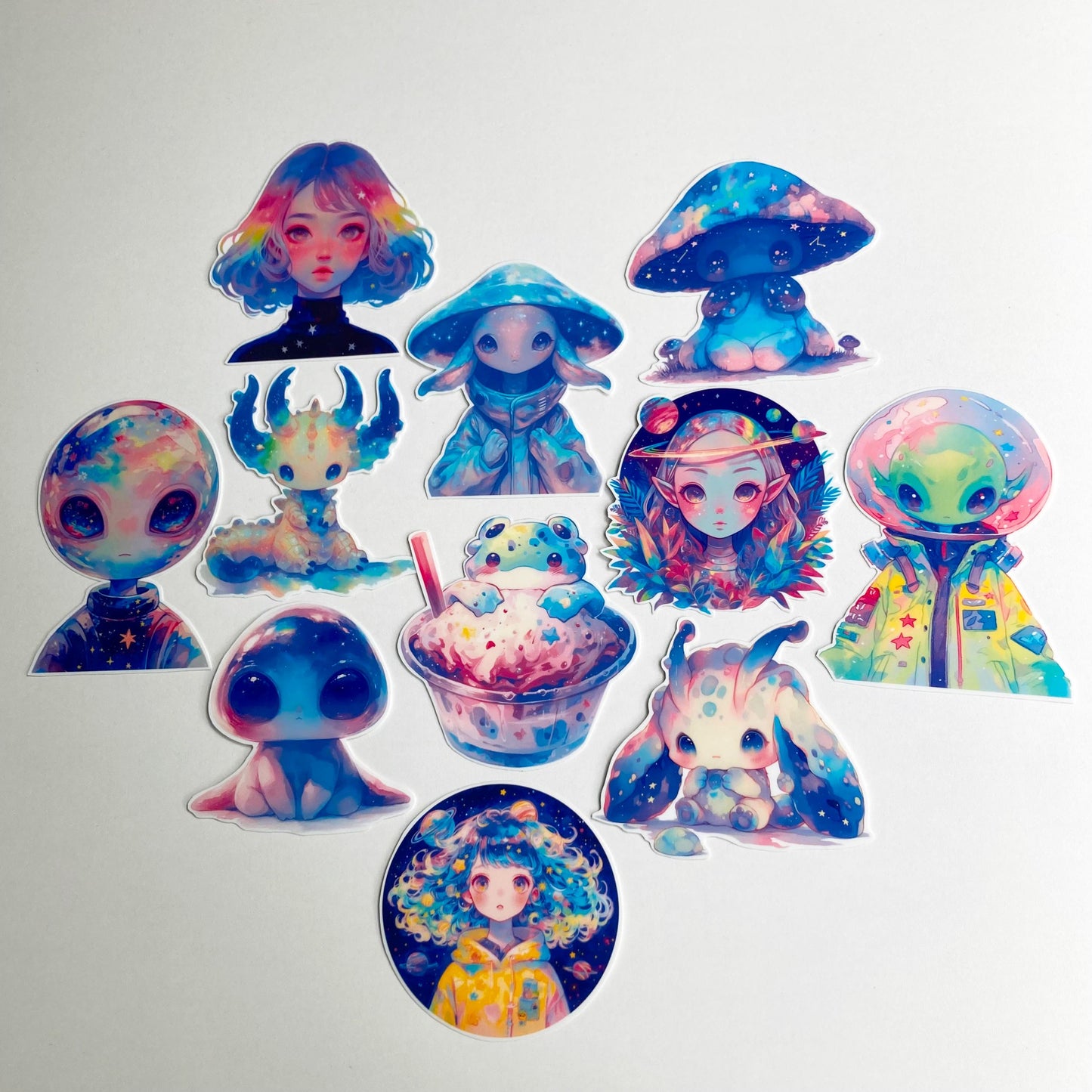 A collection of colorful stickers featuring aliens and fantasy creatures with vibrant pastel and galaxy-themed designs. In the center is a frog alien, surrounded by other otherworldly figures, including mushroom-headed beings and humanoid aliens.