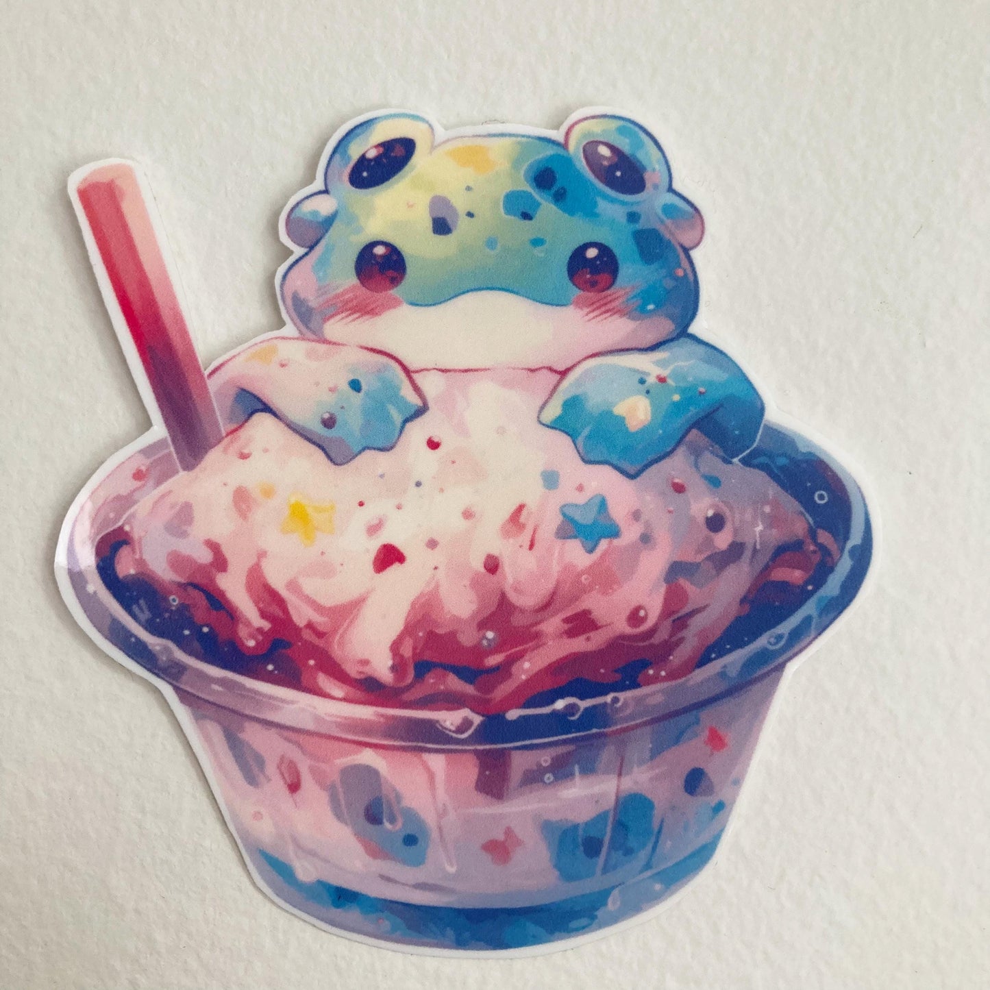 A colorful sticker of an alien frog sitting in a cup of pastel pink and blue ice cream with star-shaped sprinkles and a red-striped straw, set against a plain white background.