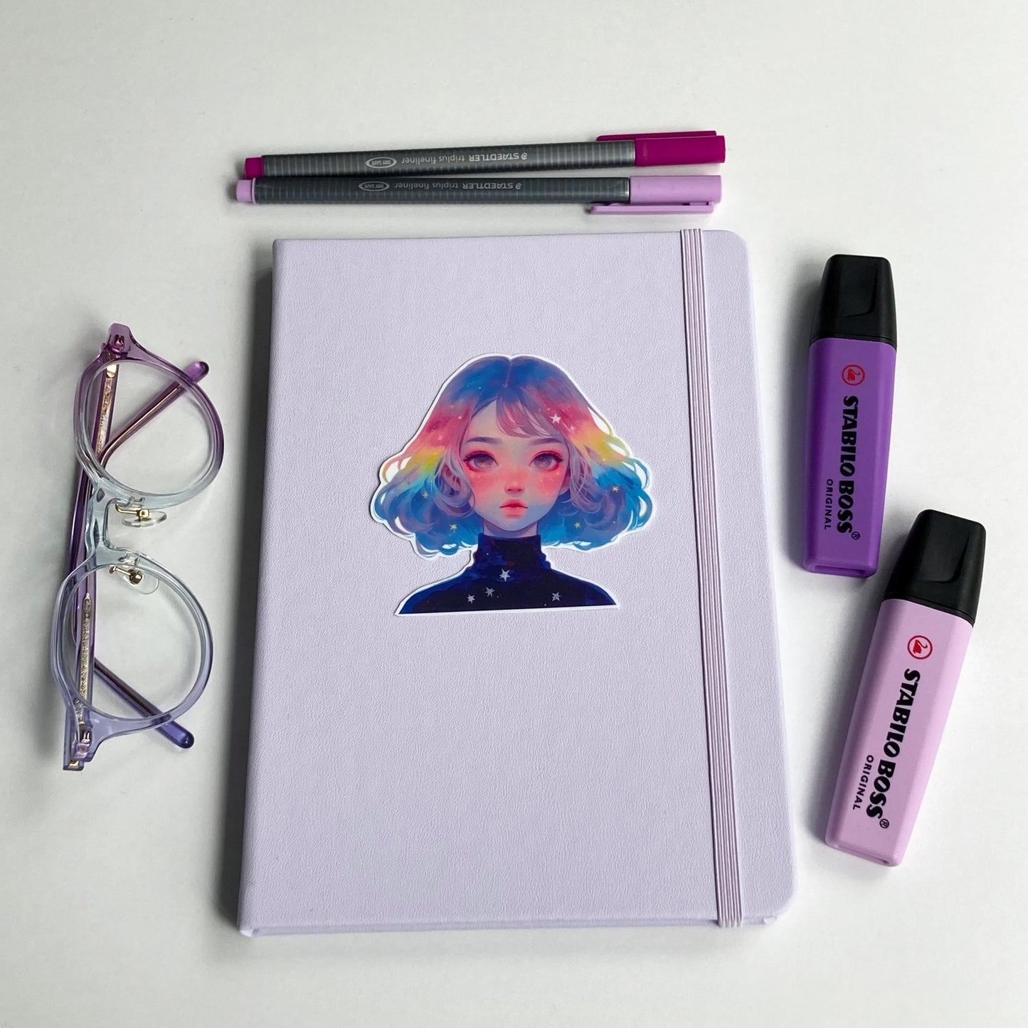 A lavender notebook with a sticker of a person with pastel rainbow hair is surrounded by purple-rimmed glasses, two highlighters, and two pens in shades of purple and pink. The items are neatly arranged on a white surface.