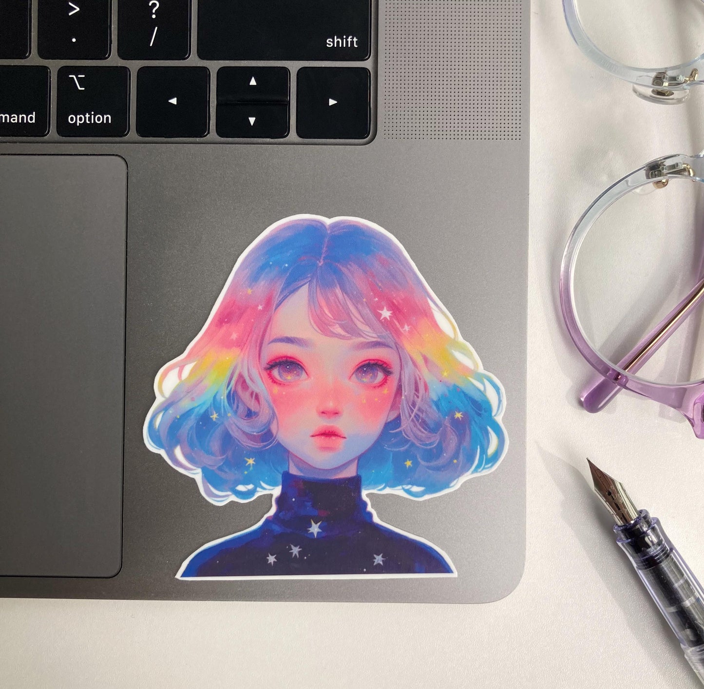 A colorful sticker of a person with pastel rainbow hair and a starry galaxy-themed shirt is placed on a laptop. Nearby are a pair of purple-rimmed glasses and a clear fountain pen.